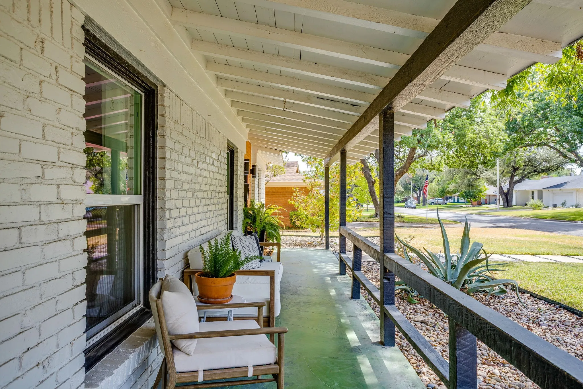 Covered front porch featuring a painted brick exterior, multiple windows, a wood-beam ceiling, and stone landscaping