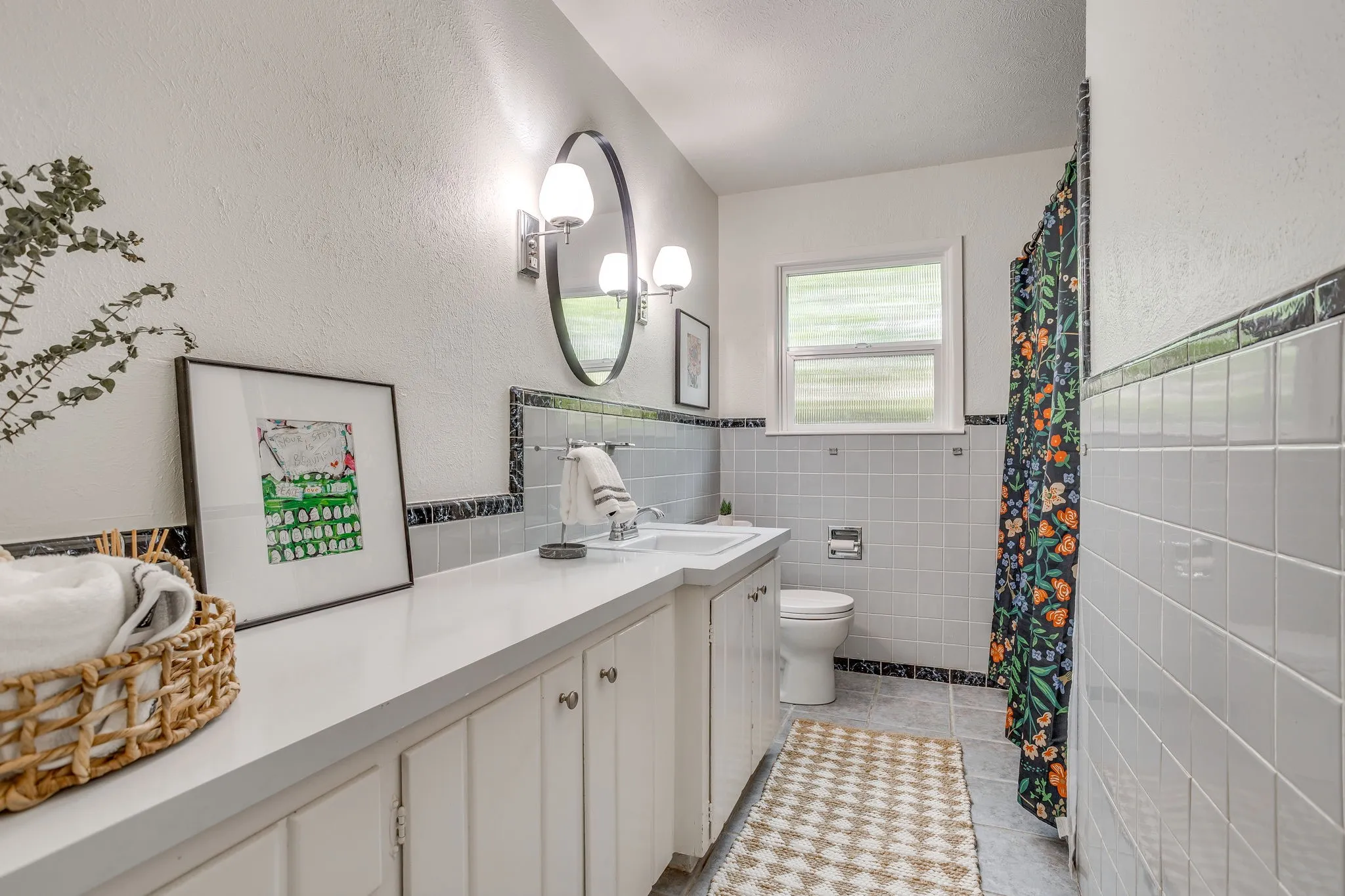 Bathroom featuring a single vanity with extensive countertop space, an oval mirror, wall-mounted light fixtures, a window, and tile-lined walls