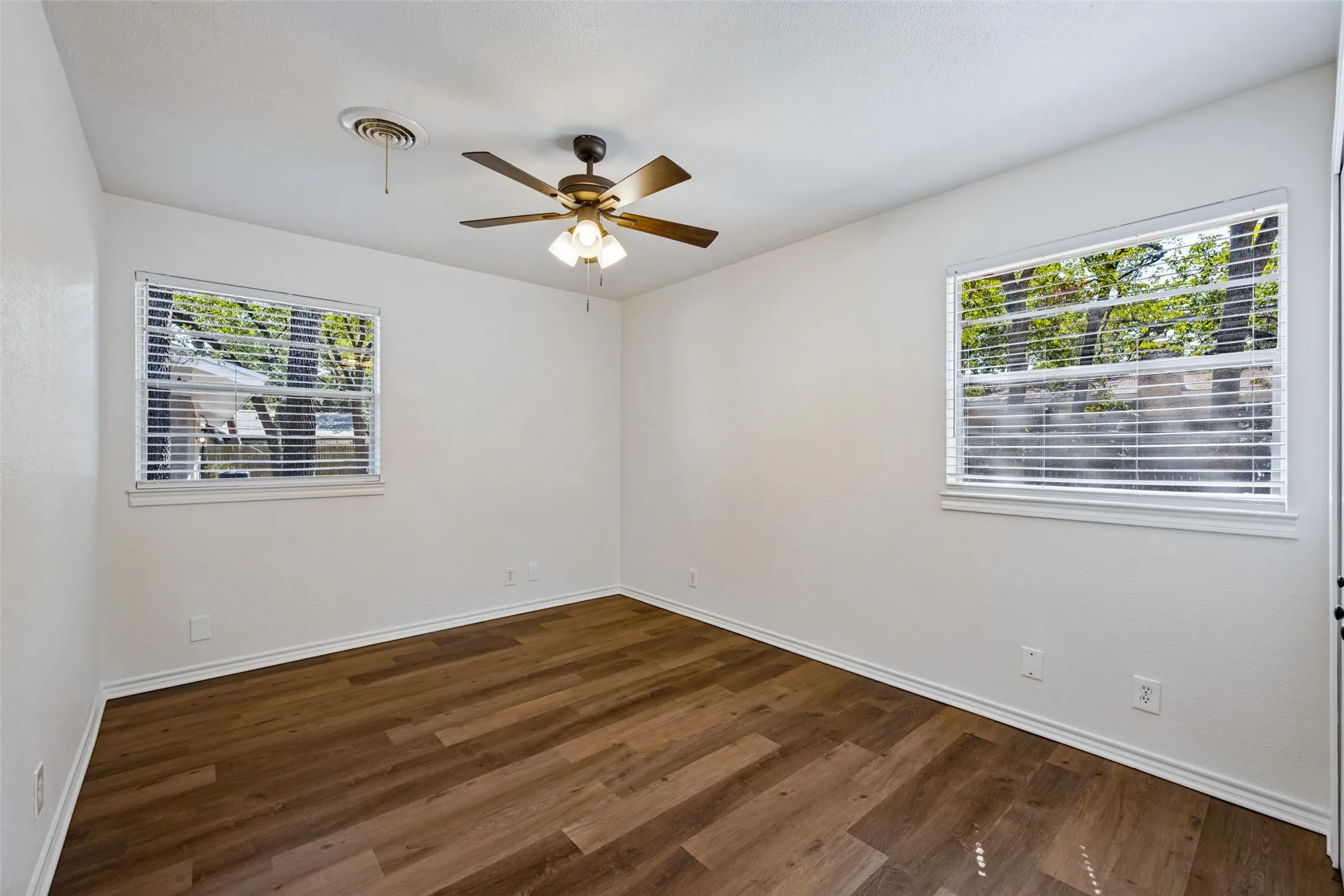Wood-finish flooring, white walls, two windows with blinds, and a ceiling fan with integrated lighting