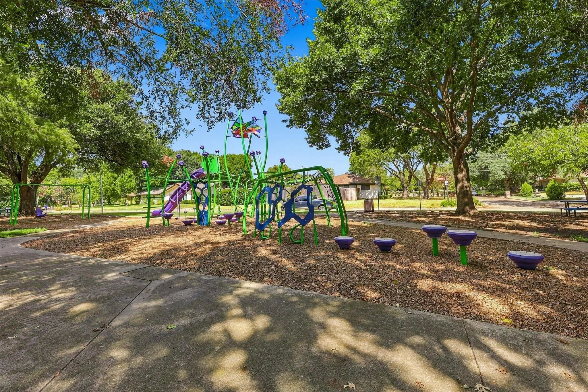 Community playground with diverse play equipment, including slides, swings, and climbing structures, set on a wood chip surface, surrounded by mature trees