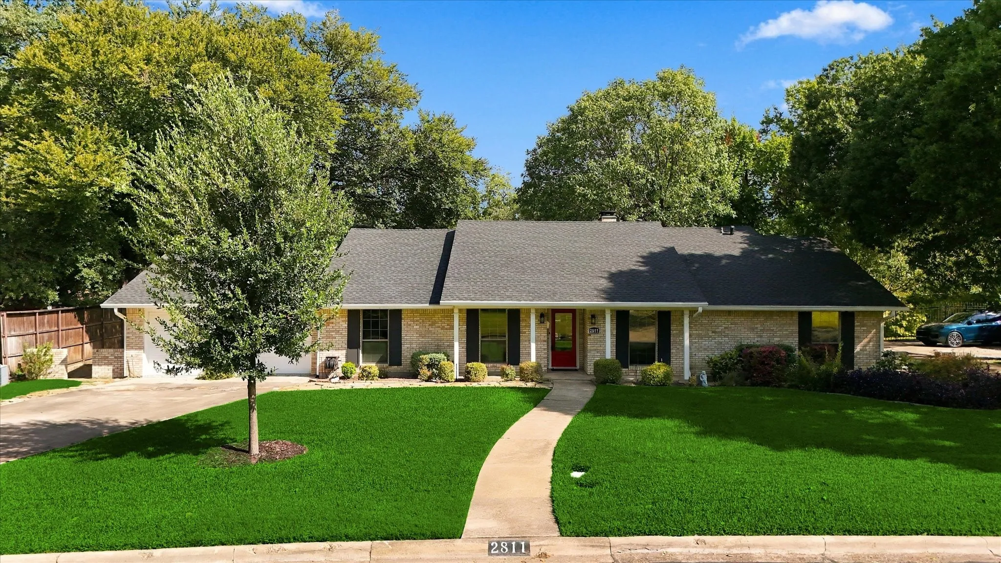 Brick facade residence with a dark shingle roof, established trees, a concrete pathway, and an attached two-car garage