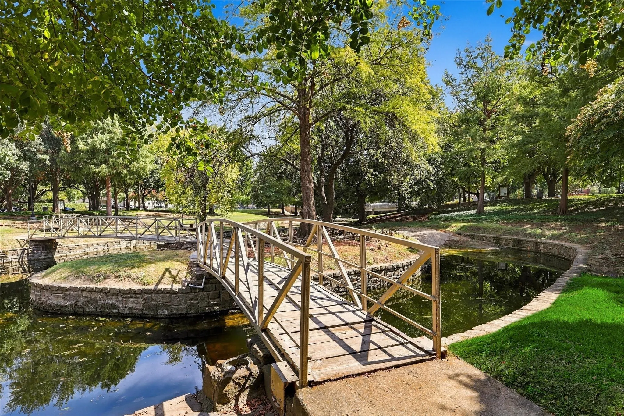 Landscaped grounds featuring a footbridge over water, surrounded by mature trees and lush greenery