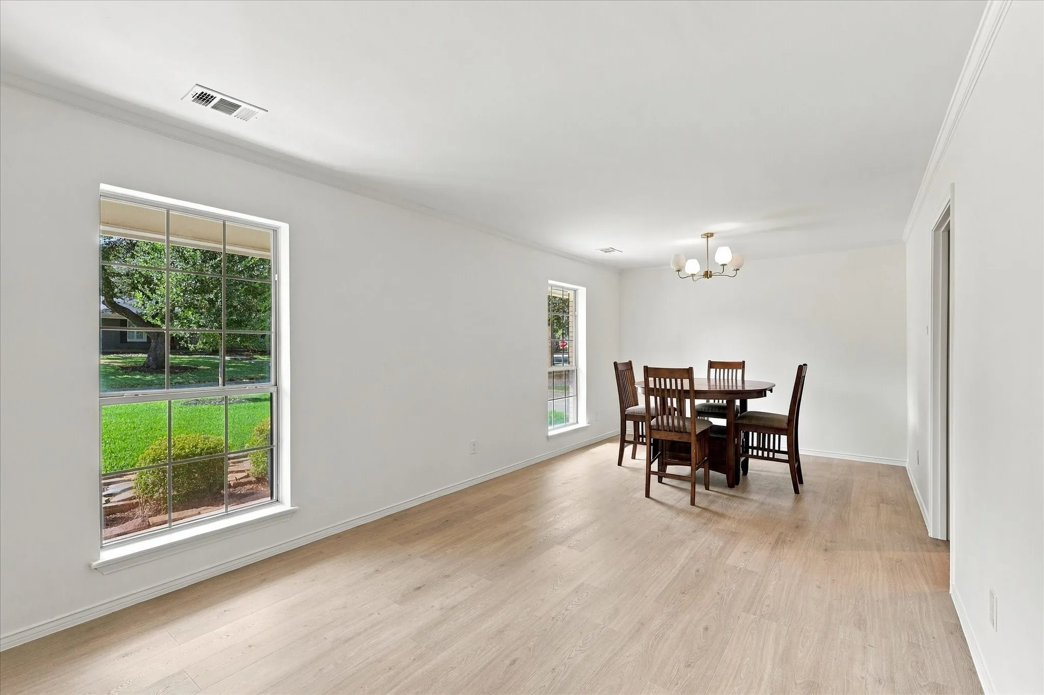 Light wood-finish flooring throughout, white walls, multiple windows, and a chandelier with frosted glass shades