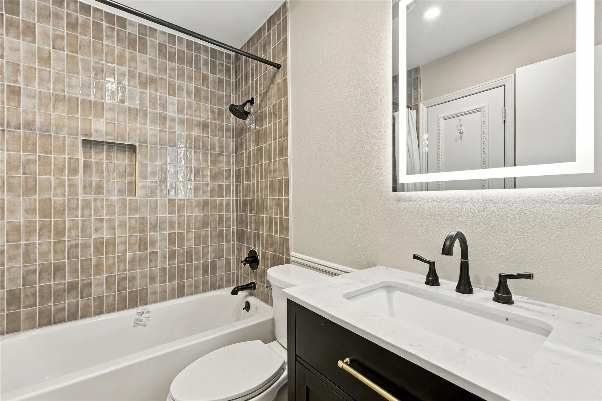 Bathroom featuring a tiled shower surround with a built-in niche, a white tub, a dark wood-finish vanity with gold-tone hardware, a white countertop, and an illuminated mirror