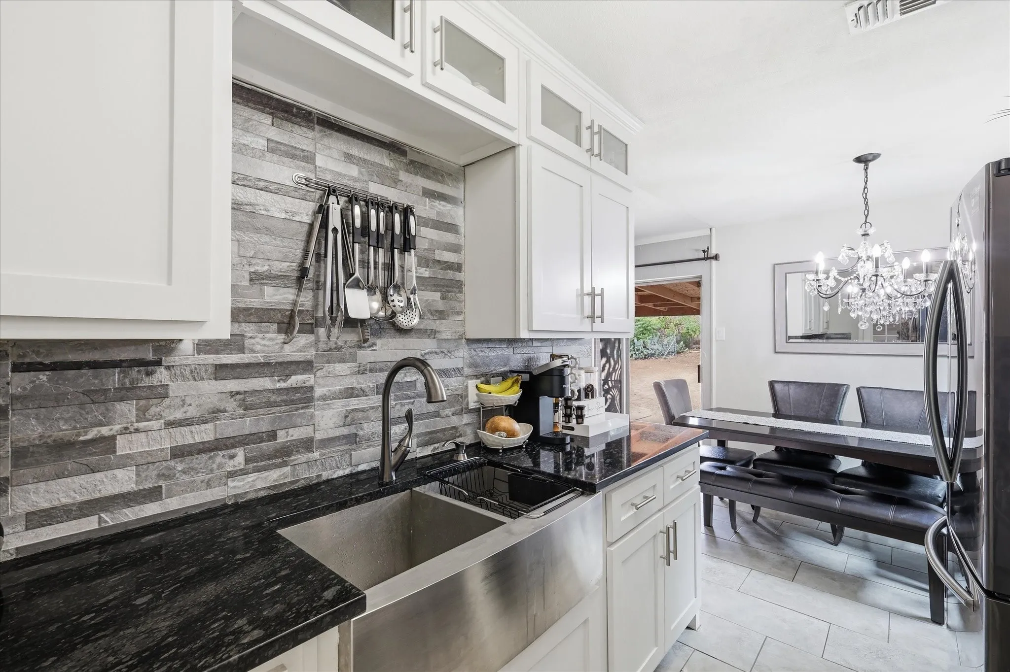 Kitchen featuring white shaker cabinetry, dark stone countertops, a stainless steel farmhouse sink, and a stacked stone-look backsplash