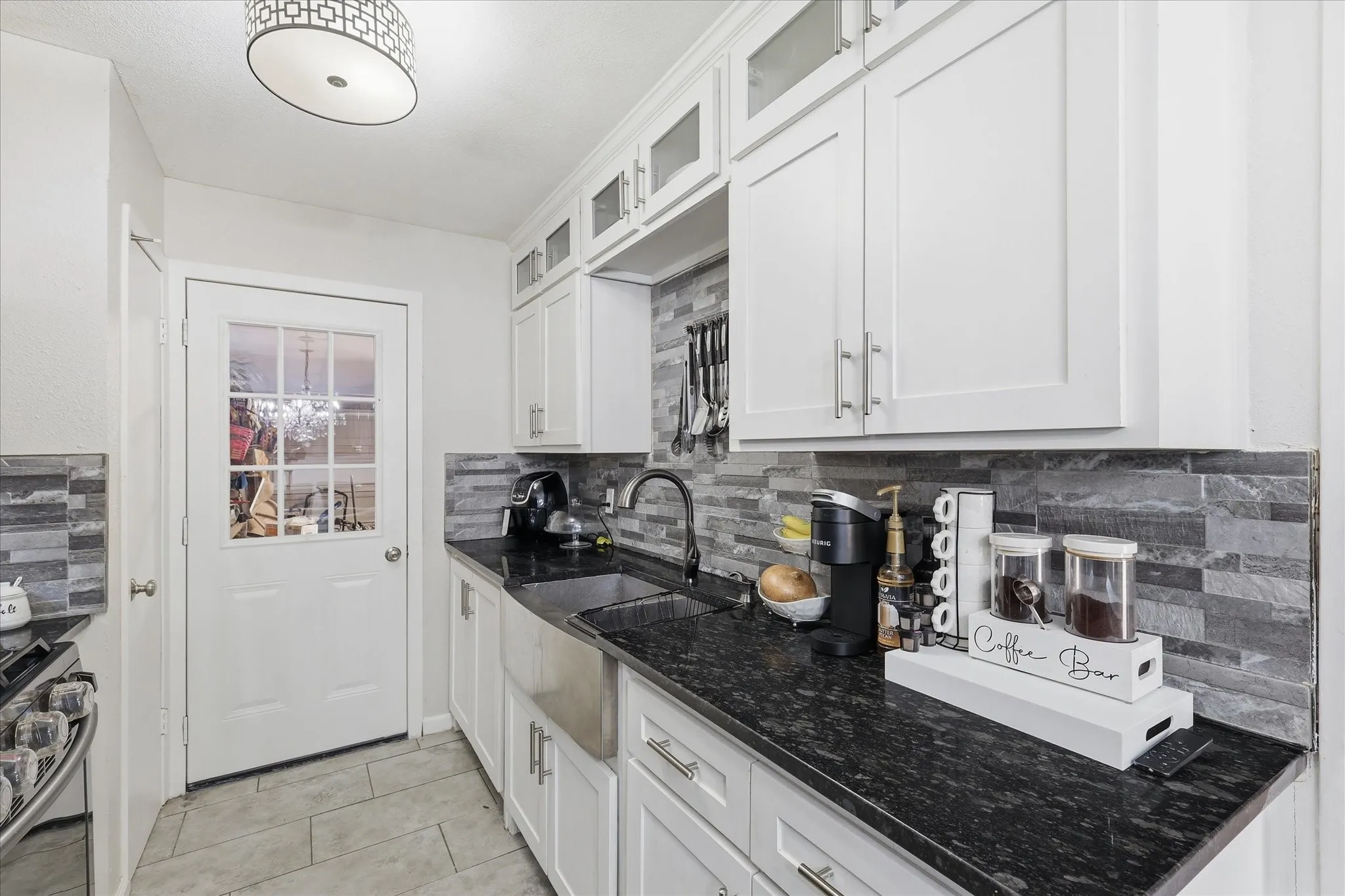 White shaker cabinetry, black stone countertops, stainless steel farmhouse sink, stacked stone backsplash, and tile flooring