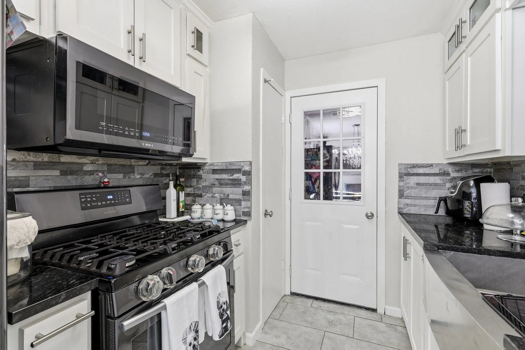 White shaker-style cabinetry with brushed nickel hardware, built-in microwave, black gas range, dark stone countertops, and a stone tile backsplash