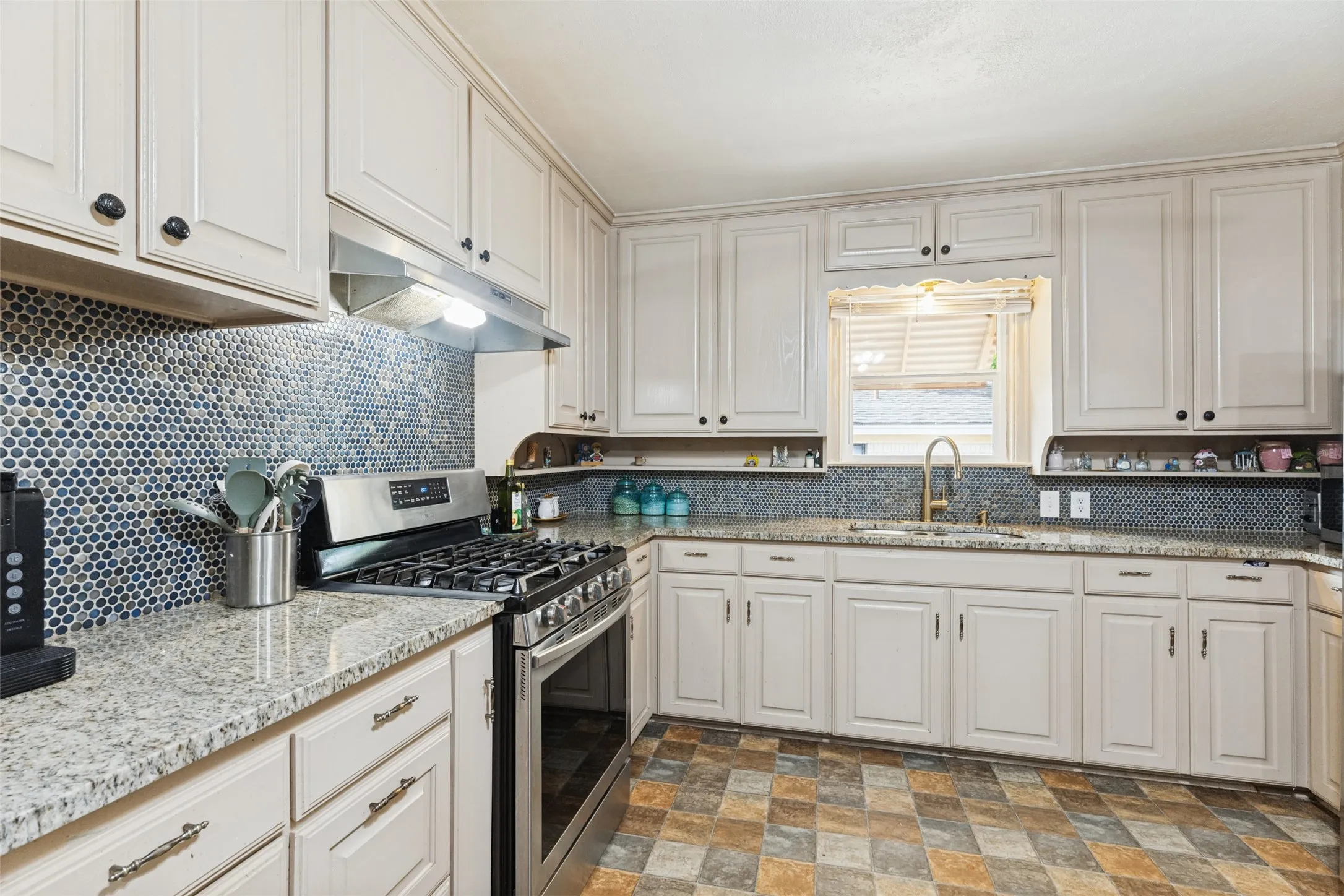 Kitchen featuring extensive light-toned cabinetry, granite-style countertops, mosaic tile backsplash, stainless steel range with hood, and a gold-finish faucet