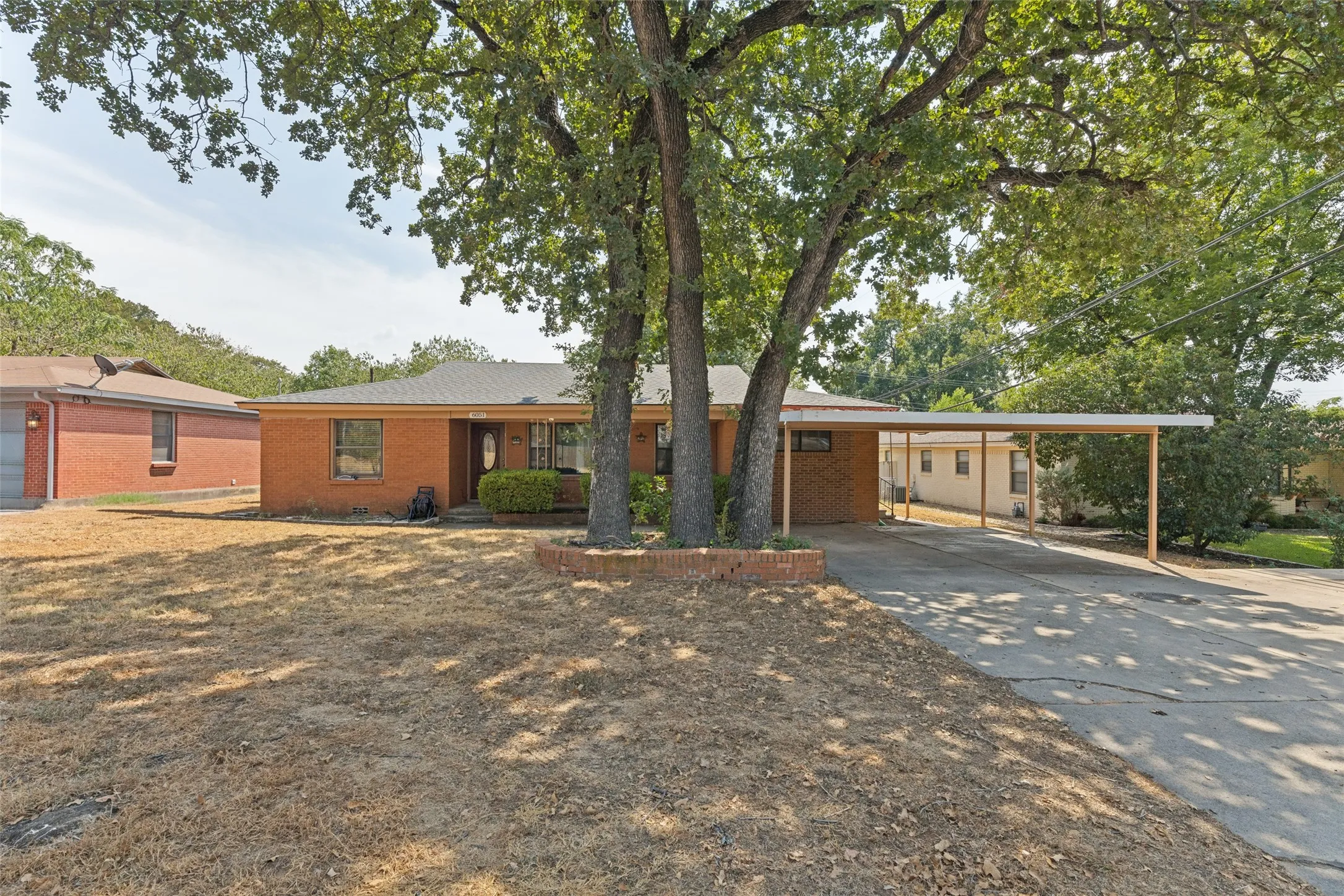 Brick exterior with a covered carport, mature trees, and a low brick planter