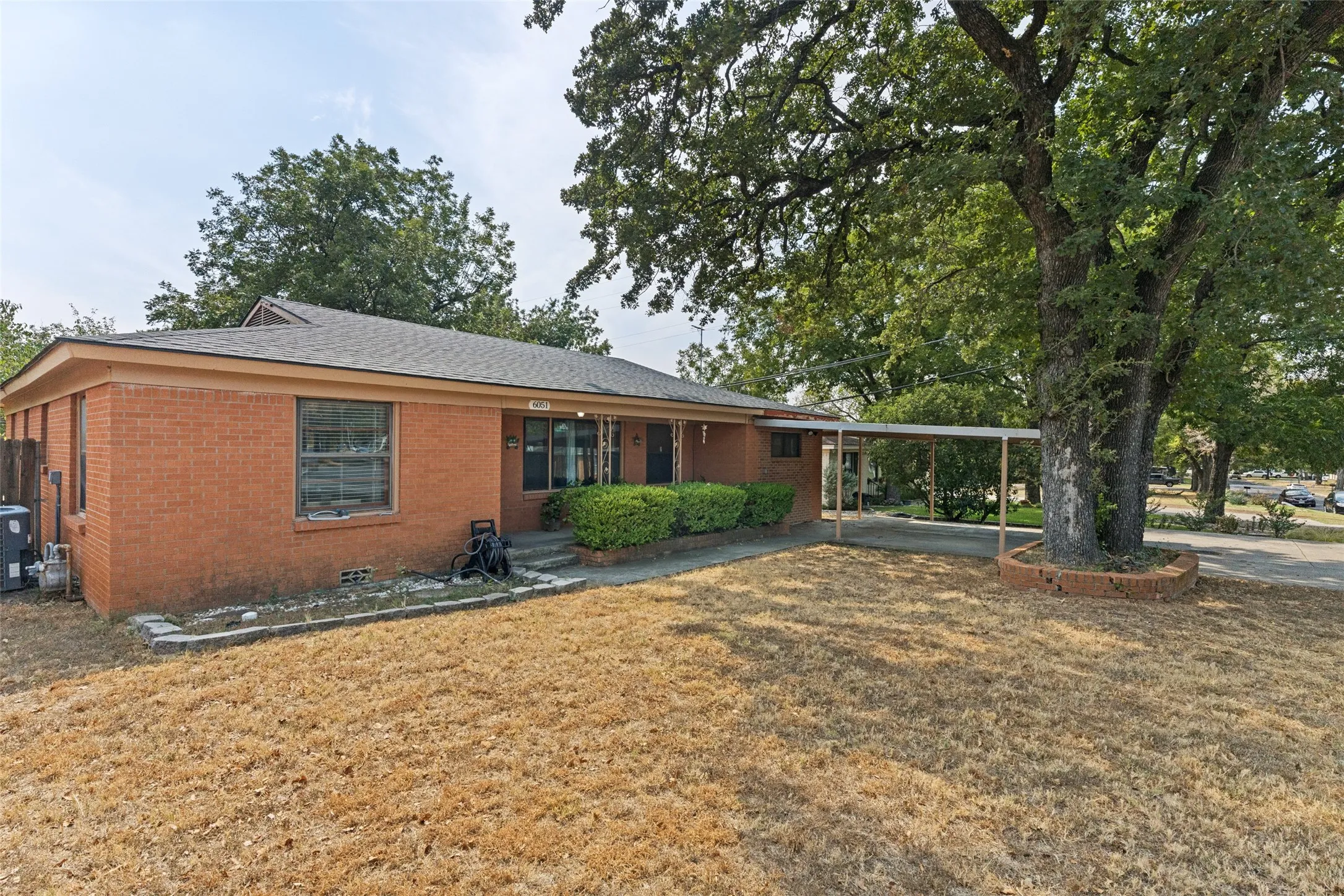 Brick exterior with a covered carport, mature shade tree, and established shrubbery