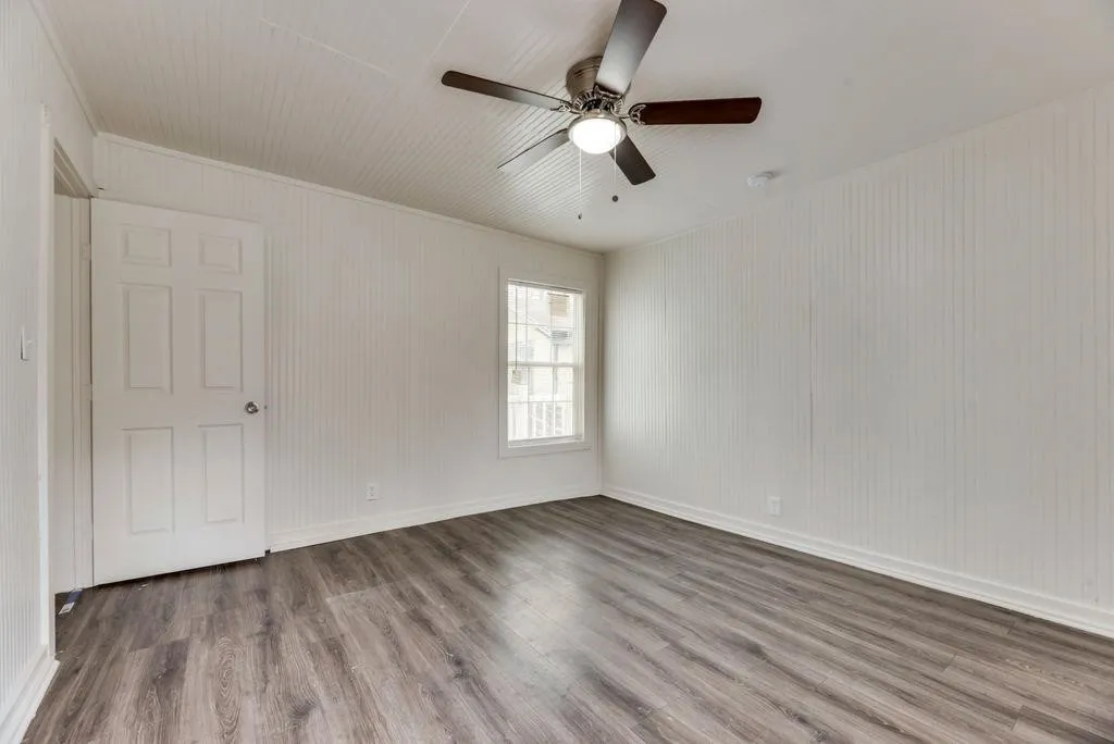 Wood-finish flooring, beadboard wall paneling, white trim, ceiling fan with integrated lighting, and a multi-pane window