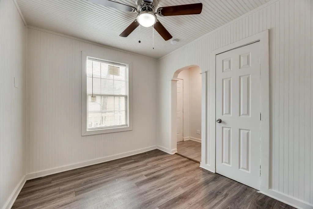 Wood-finish flooring, beadboard wall paneling, a single window, a ceiling fan with integrated lighting, and an arched doorway