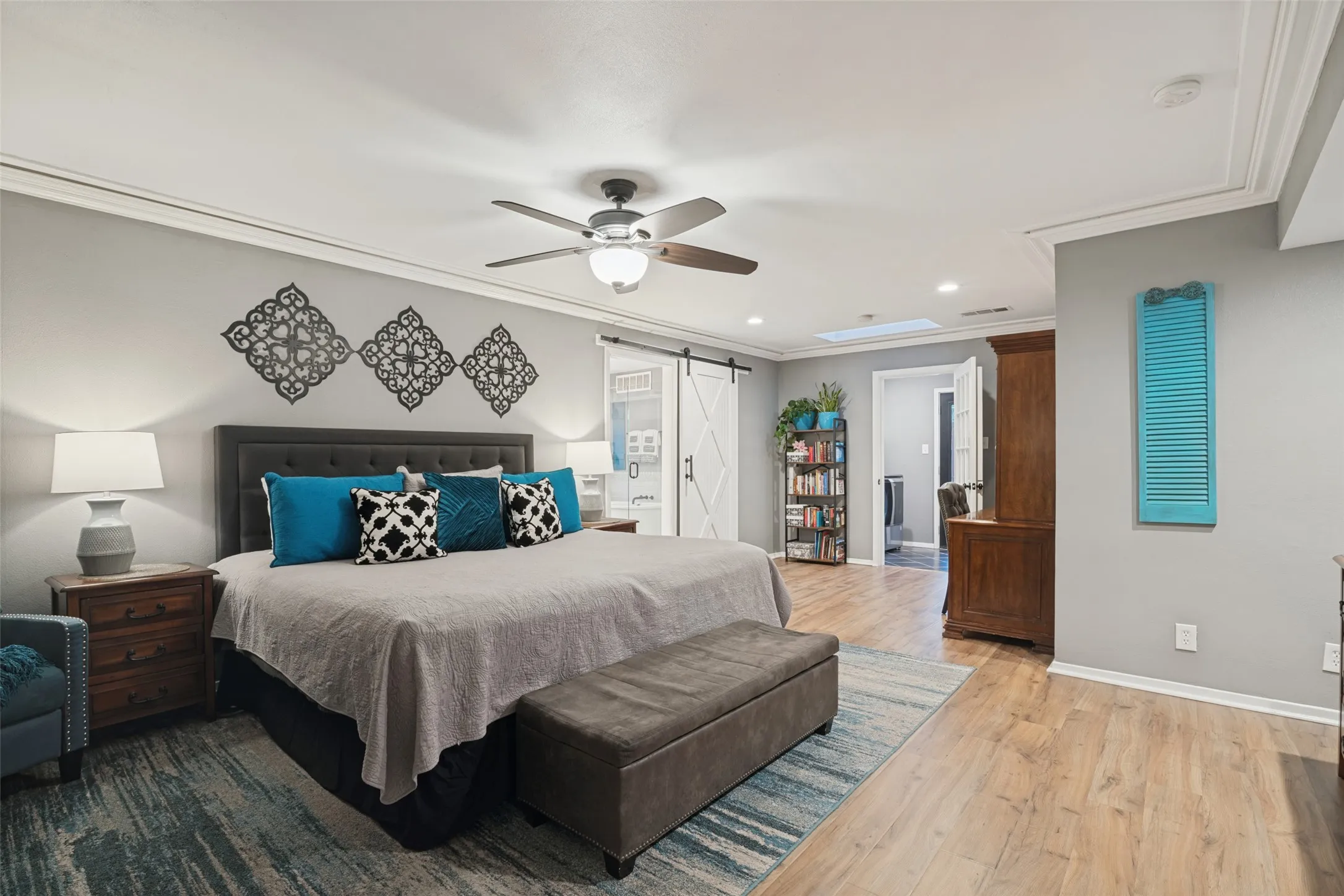 The spacious primary bedroom includes room for a seating area, convenient access to the utility room, a sky light, crown molding, an upgraded ceiing fan, and a custom barn-style door leading to the en-suite primary bath.