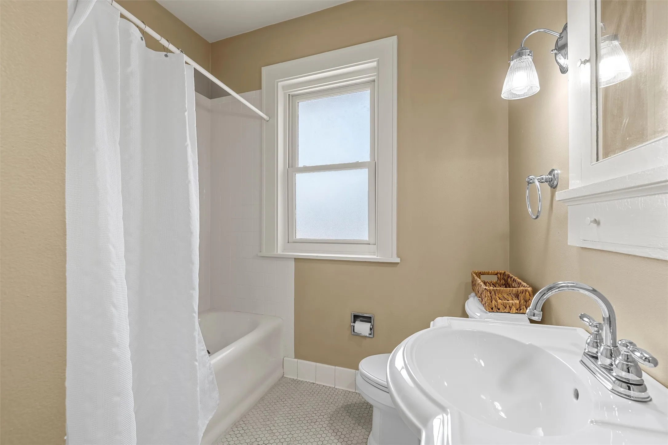 Built-in bathtub with shower, pedestal sink with chrome faucet, window with white trim, wall-mounted vanity light, and hexagonal floor tiles