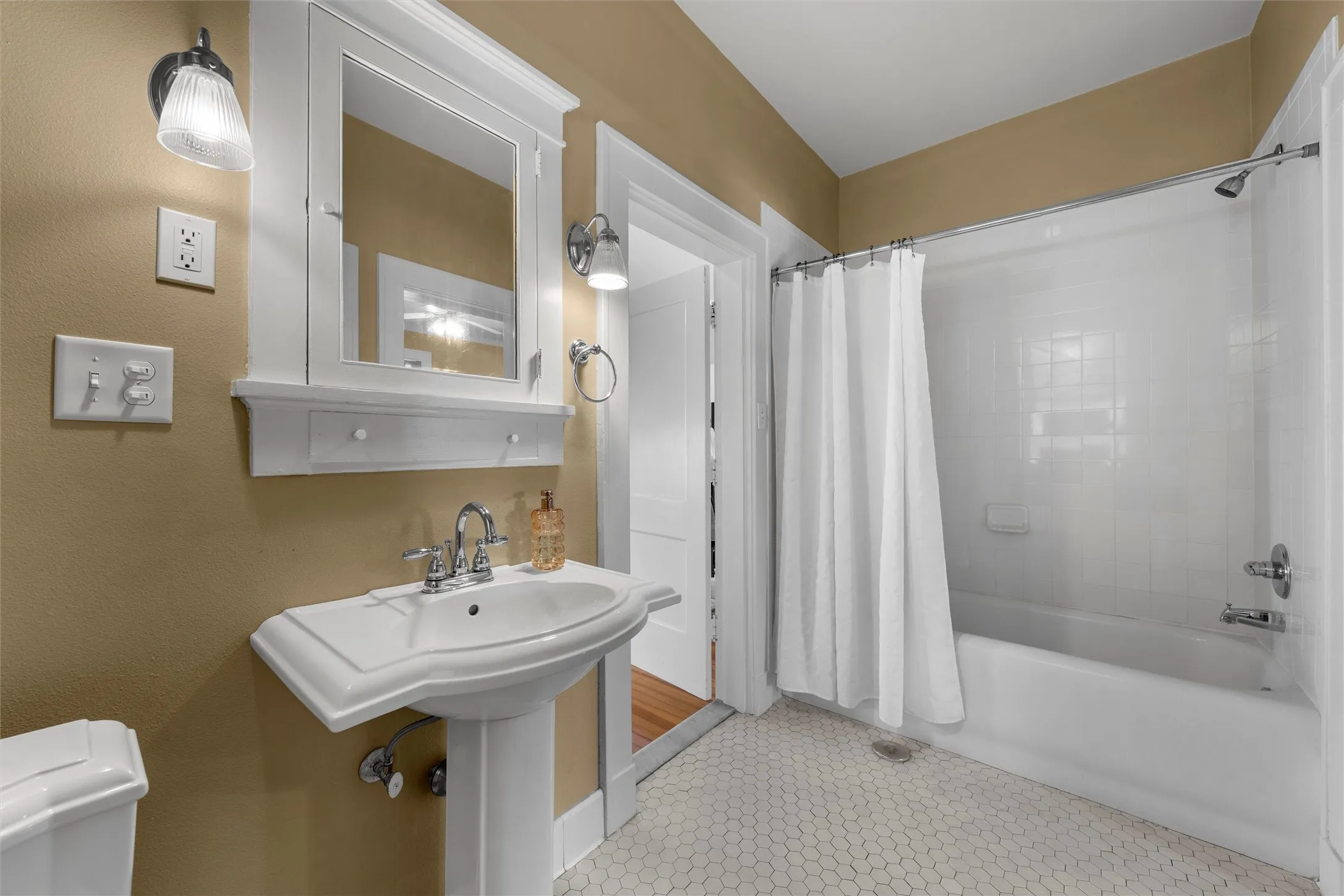 Pedestal sink with chrome faucet, framed medicine cabinet, wall-mounted light fixtures, white hexagon tile flooring, and a shower-over-tub combination