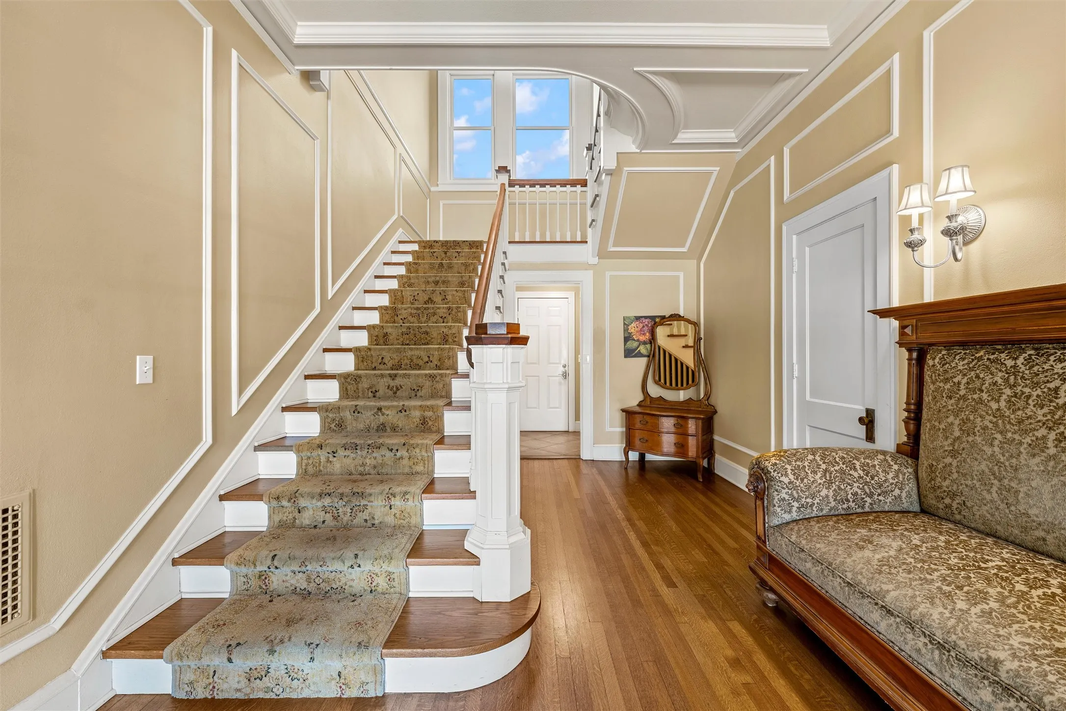 Hardwood flooring, custom wall molding, grand staircase with carpet runner, wall sconce lighting, and high ceilings