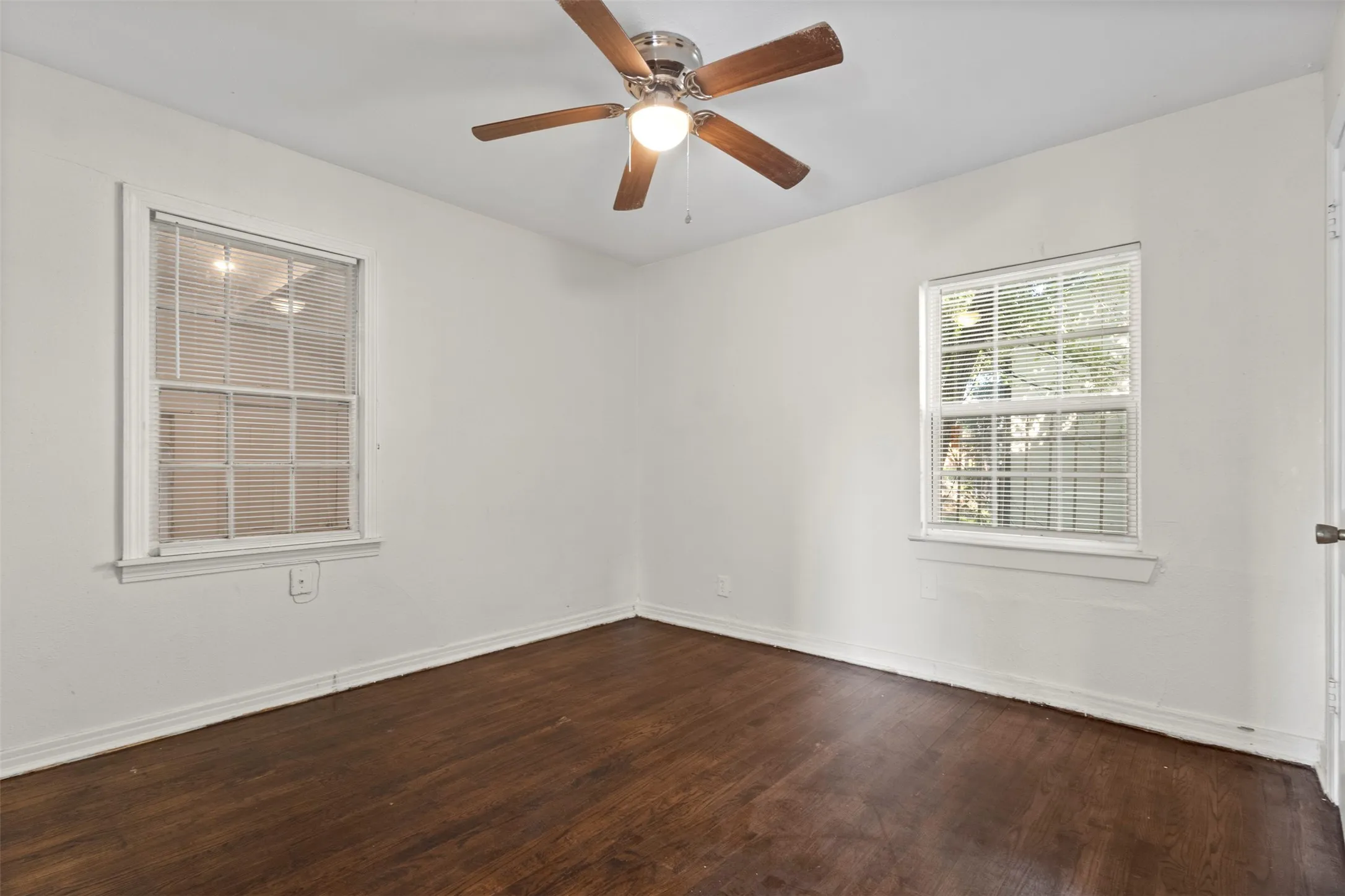 Wood-finish flooring, white walls, two windows with blinds, and a ceiling fan with light fixture