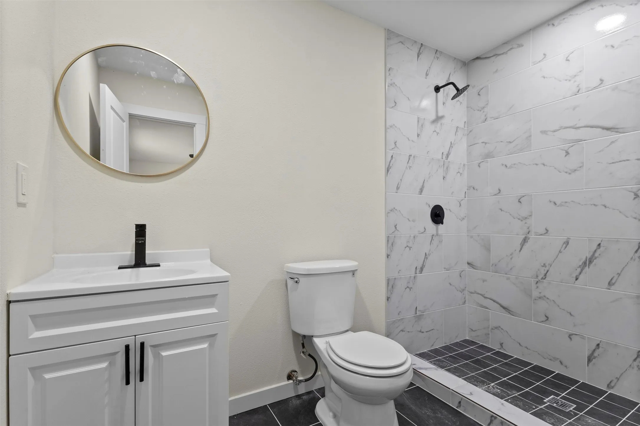 White vanity with matte black faucet, round wall mirror, and white shower with dark tile flooring