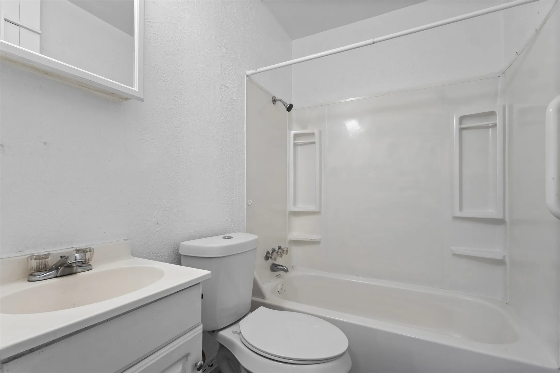 White vanity with integrated sink, chrome faucet, white toilet, and a white tub/shower combination with built-in shelving