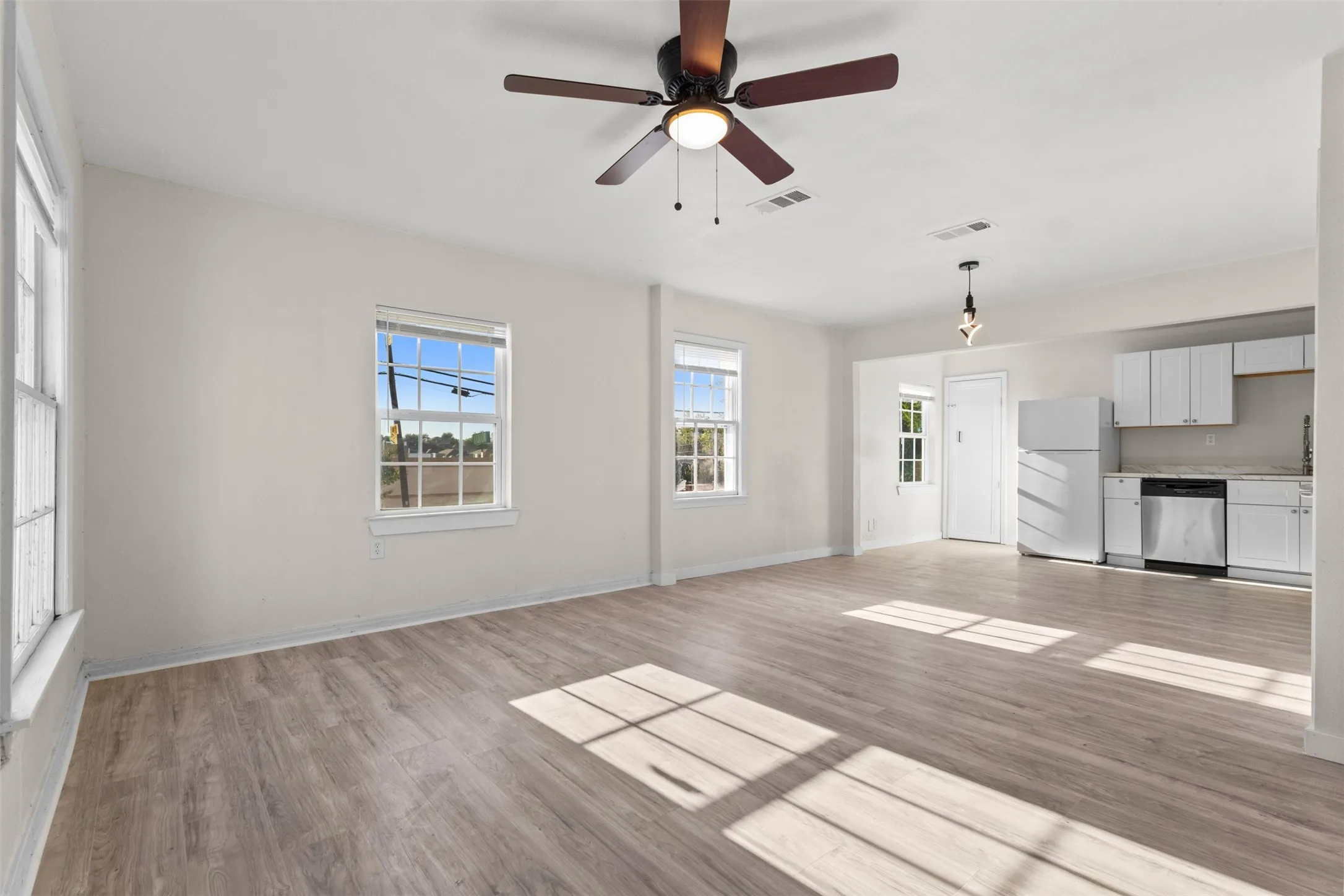 Open-concept living area with wood-finish flooring, two large windows, a ceiling fan, and a kitchen featuring white cabinetry and stainless steel appliances