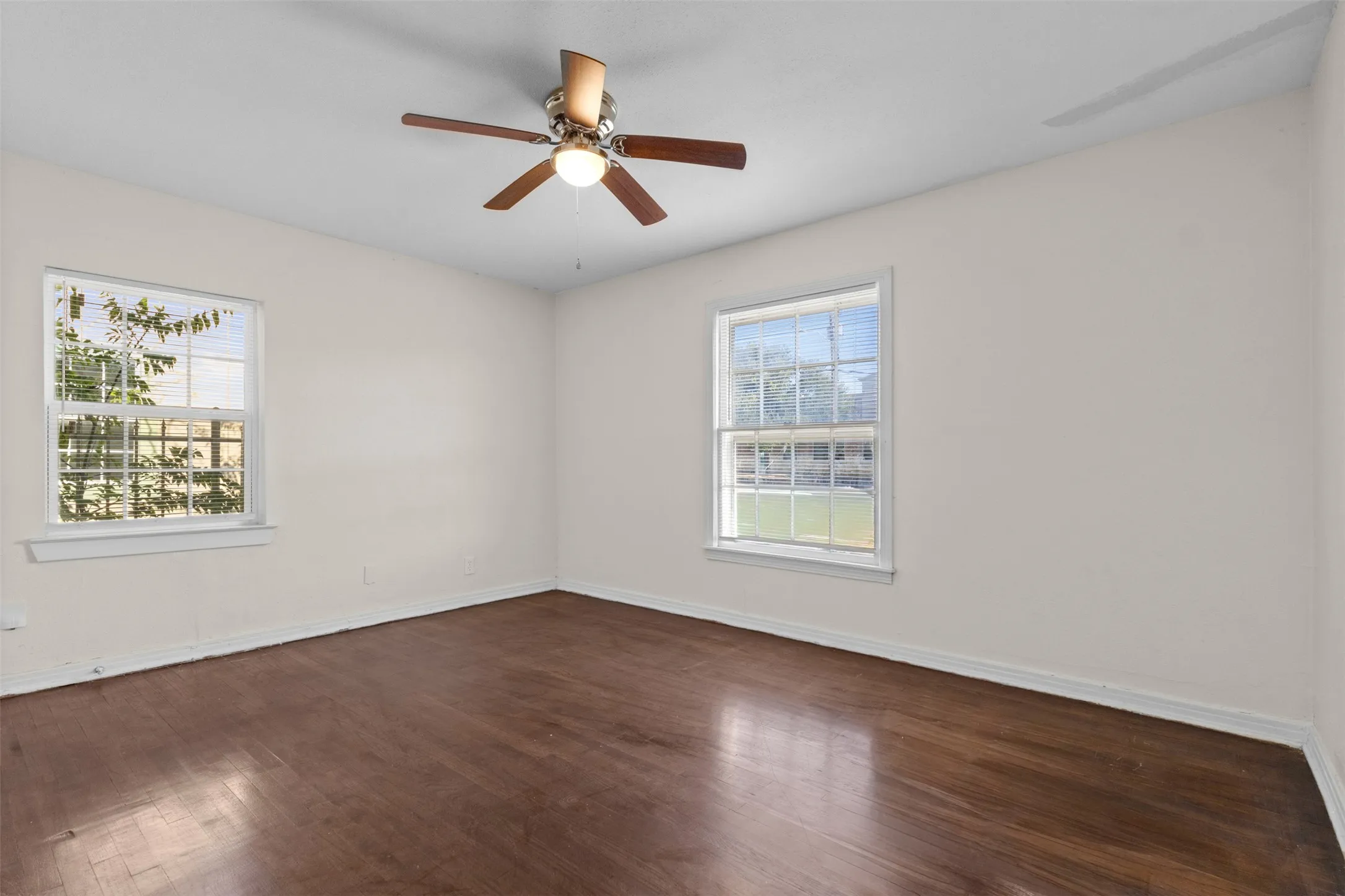 Expansive room with wood-finish flooring, two large windows, a ceiling fan, and neutral painted walls