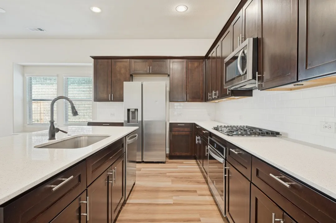 Kitchen featuring dark wood-finish cabinetry, light-toned countertops, stainless steel appliances, a center island with sink, and wood-finish flooring