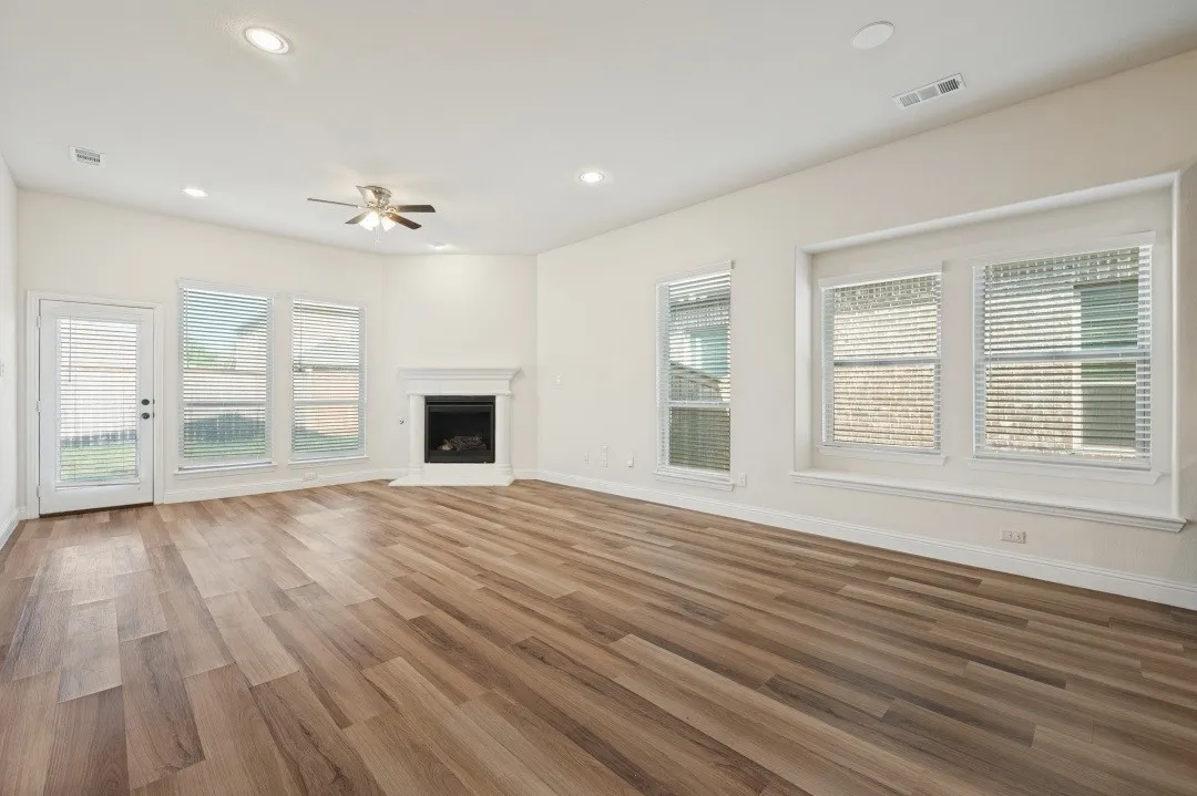 Spacious living area featuring wood-finish flooring, a corner fireplace with a classic mantel, multiple windows with blinds, a ceiling fan, and recessed lighting