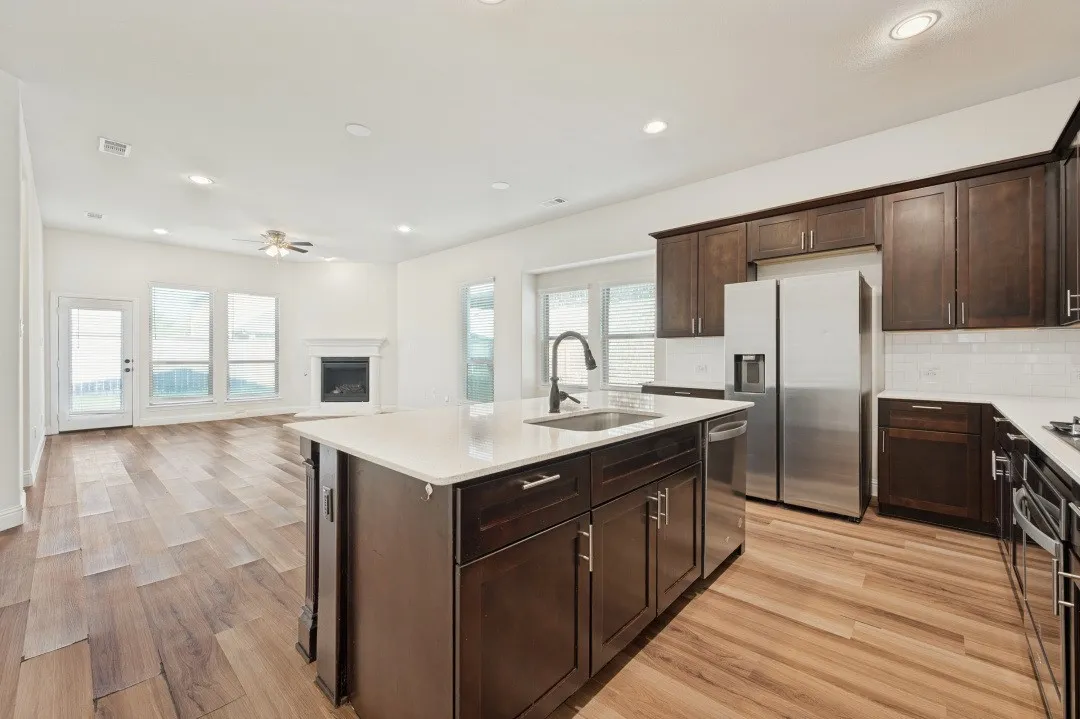 Open-concept living area featuring wood-finish flooring, a central kitchen island with a built-in sink, stainless steel appliances, and a fireplace with a mantel