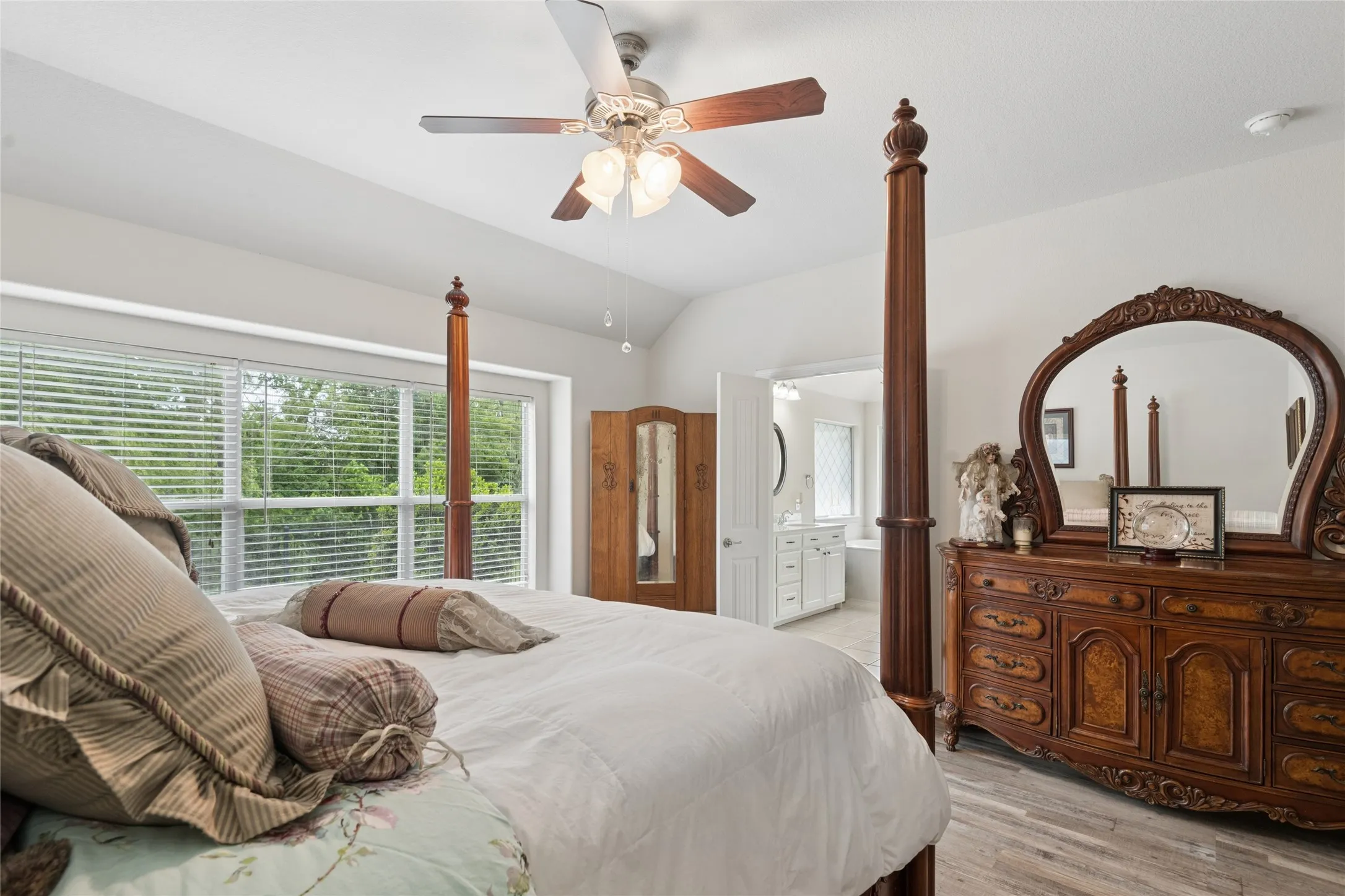 Vaulted ceiling with a ceiling fan, large window with blinds, wood-finish flooring, and an adjoining bathroom with a vanity