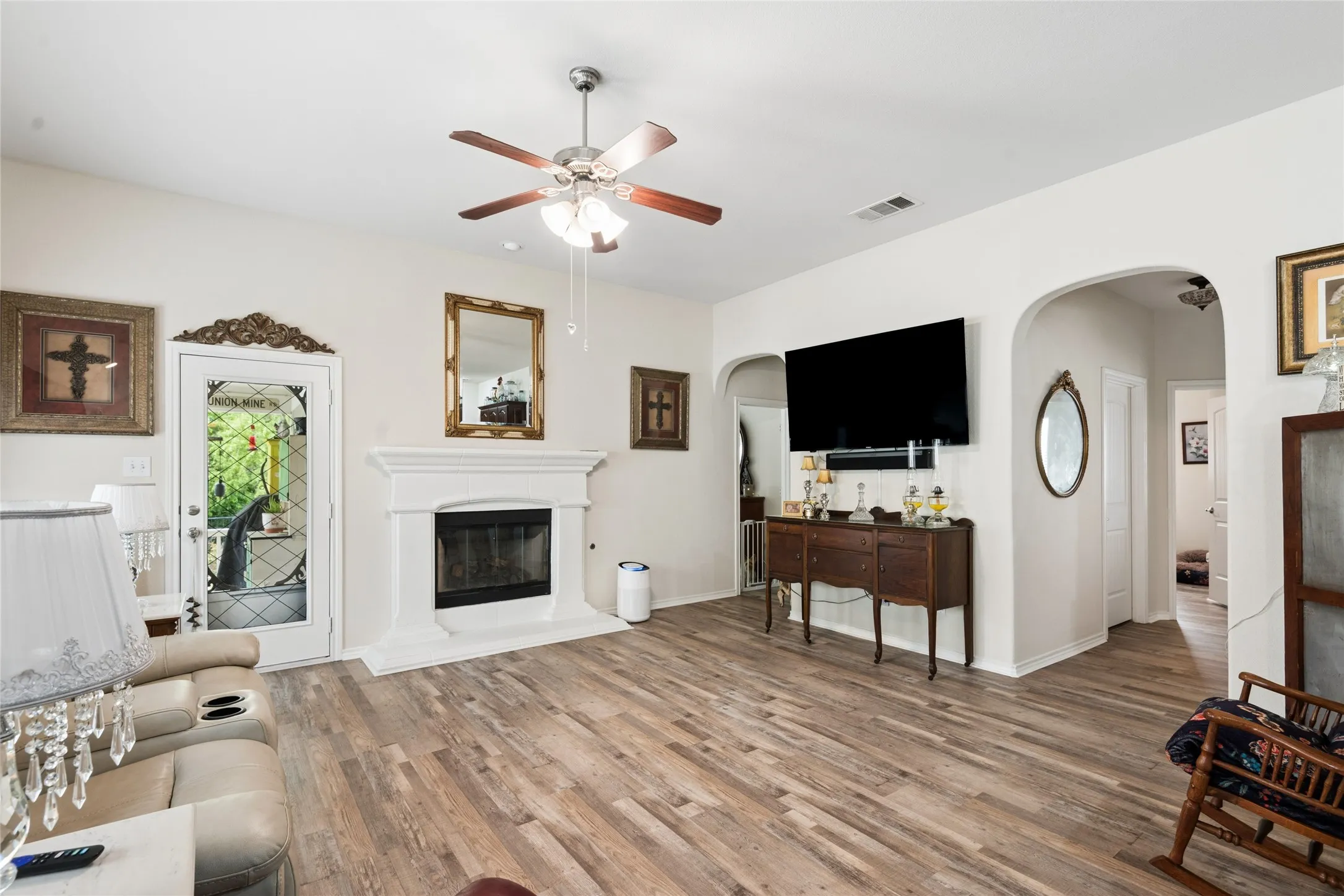 Wood-finish flooring, white fireplace with hearth, white walls, ceiling fan with light fixture, and arched doorways