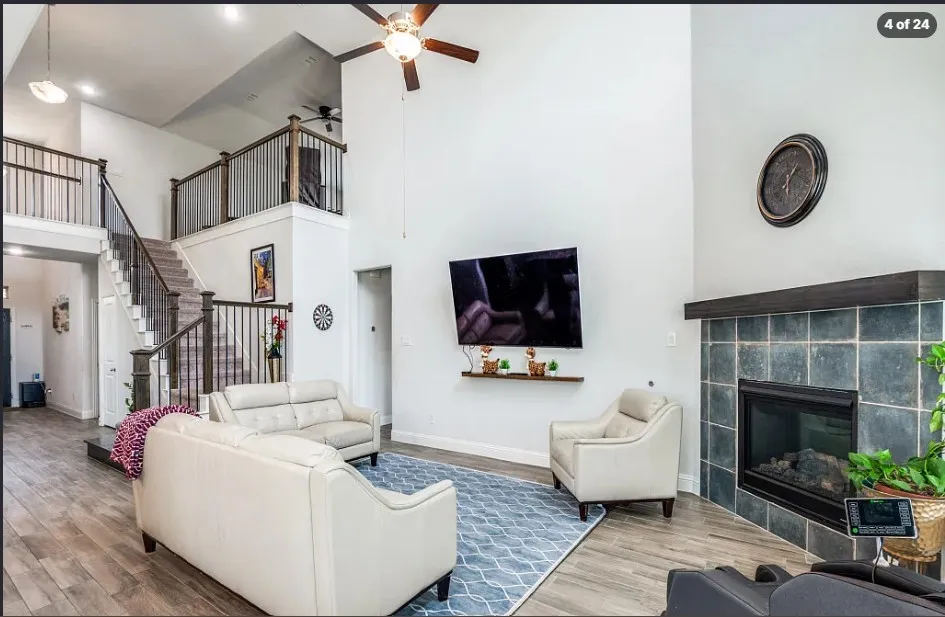 Vaulted ceiling, wood-finish flooring, two-story stone-tiled fireplace with hearth, ceiling fan, and multi-level staircase with dark wood and metal railings