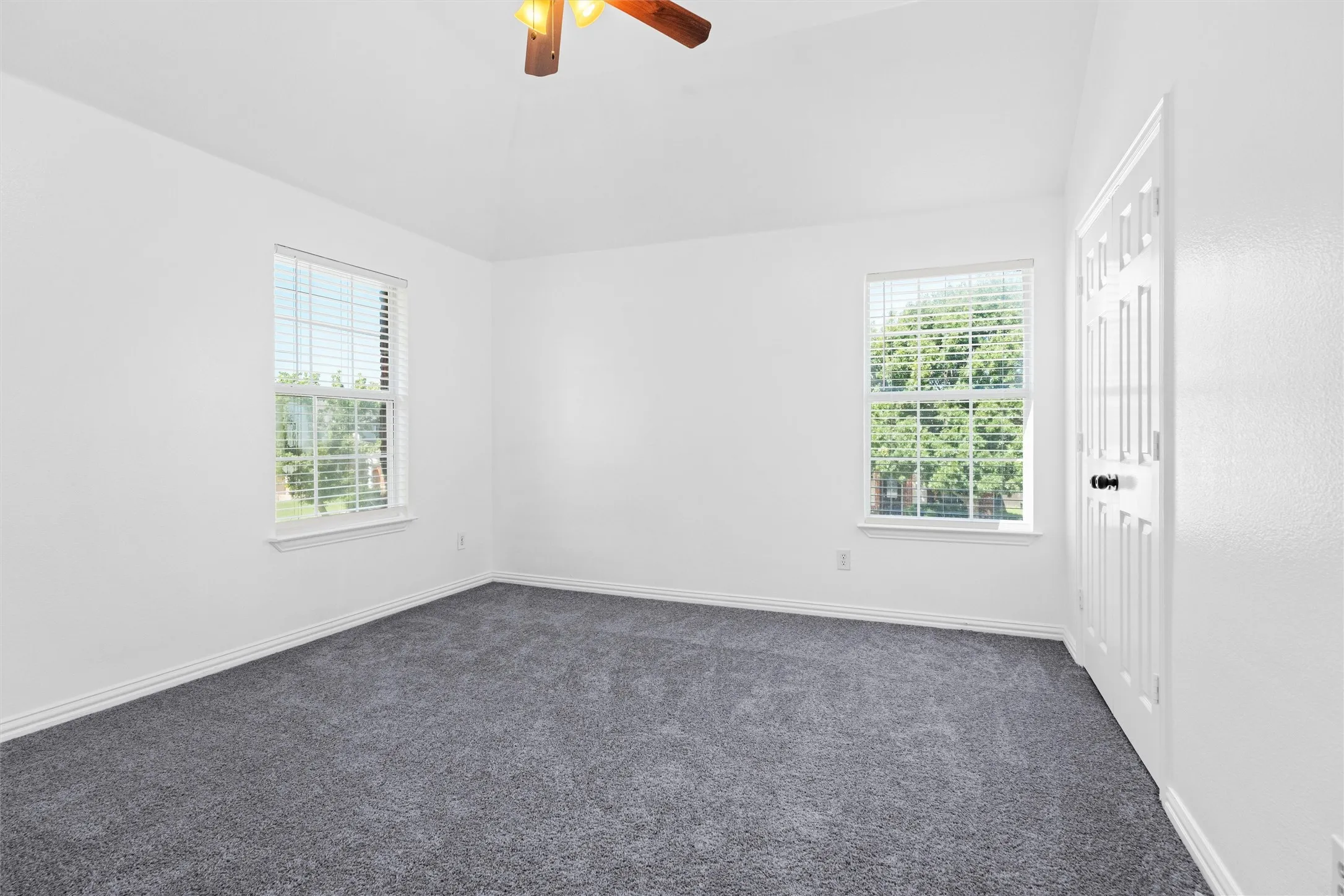 Plush gray carpeting, white walls, two window openings, and a ceiling fan with integrated lighting