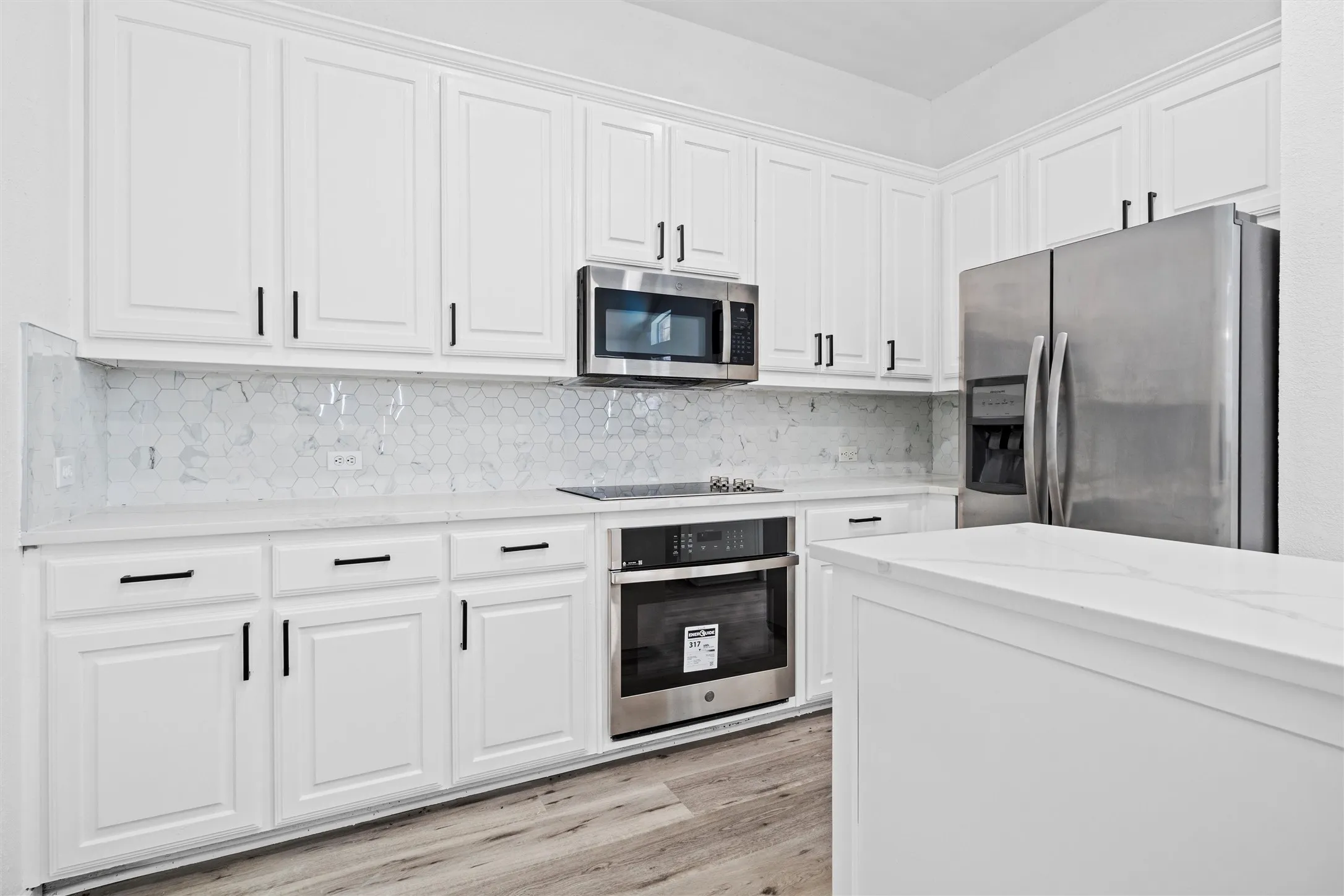 White shaker-style cabinetry with matte black hardware, stainless steel appliances, patterned tile backsplash, light-toned countertops, and wood-finish flooring