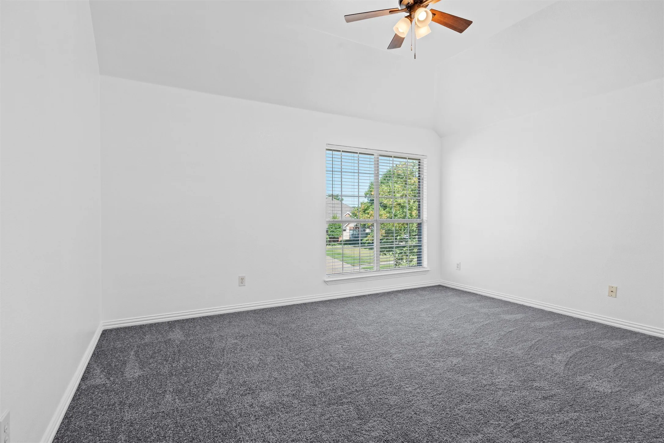 Vaulted ceiling, ceiling fan, large window with blinds, and neutral grey carpet