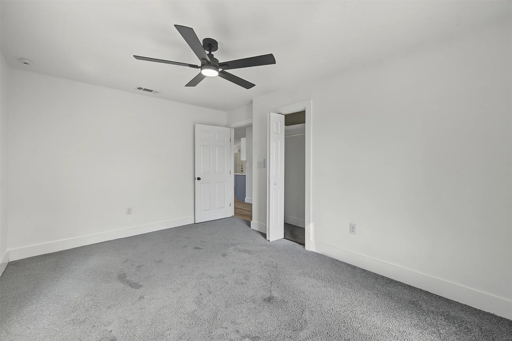 Spacious room featuring light gray carpeting, a contemporary ceiling fan with lighting, white painted walls, and a built-in closet with bi-fold doors