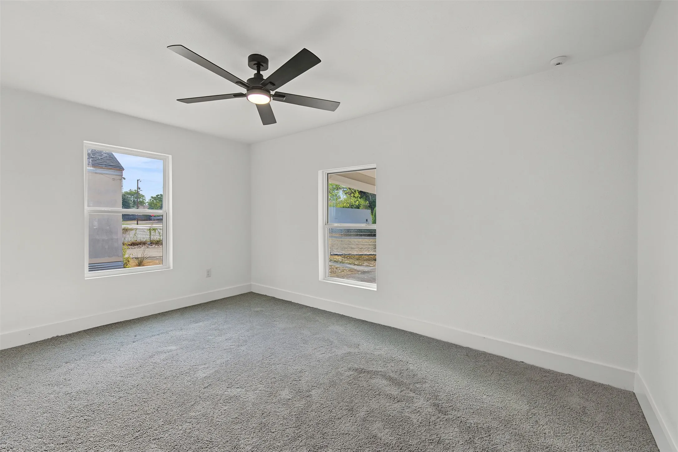 White walls with two large windows, gray carpet, and a ceiling fan with integrated lighting