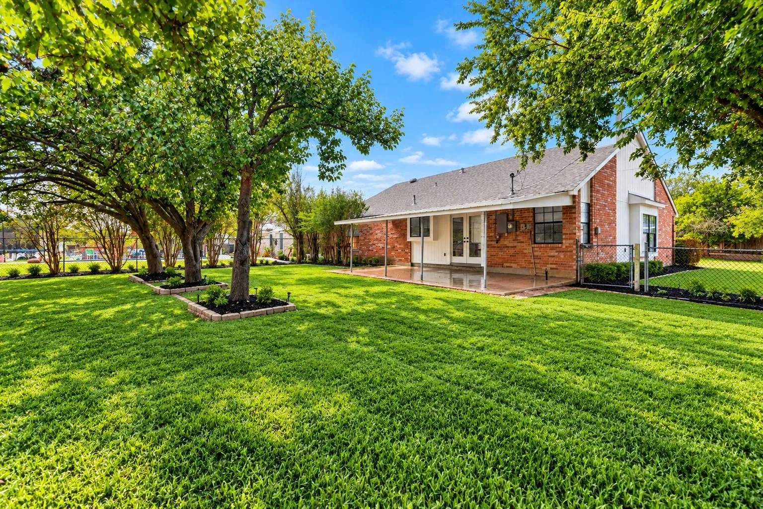 Expansive backyard featuring a manicured lawn, mature trees with mulched beds, a covered patio, and a chain-link fence
