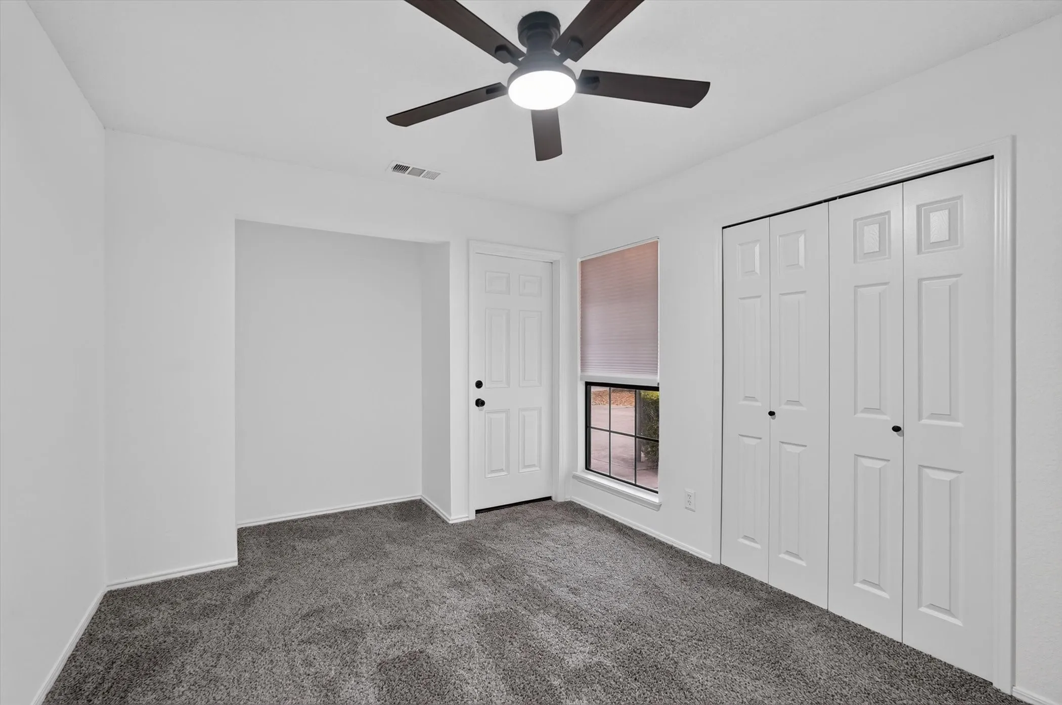 Neutral white wall finishes, plush grey carpeting, ceiling fan with light fixture, paneled bifold closet doors, and a window with roller shade