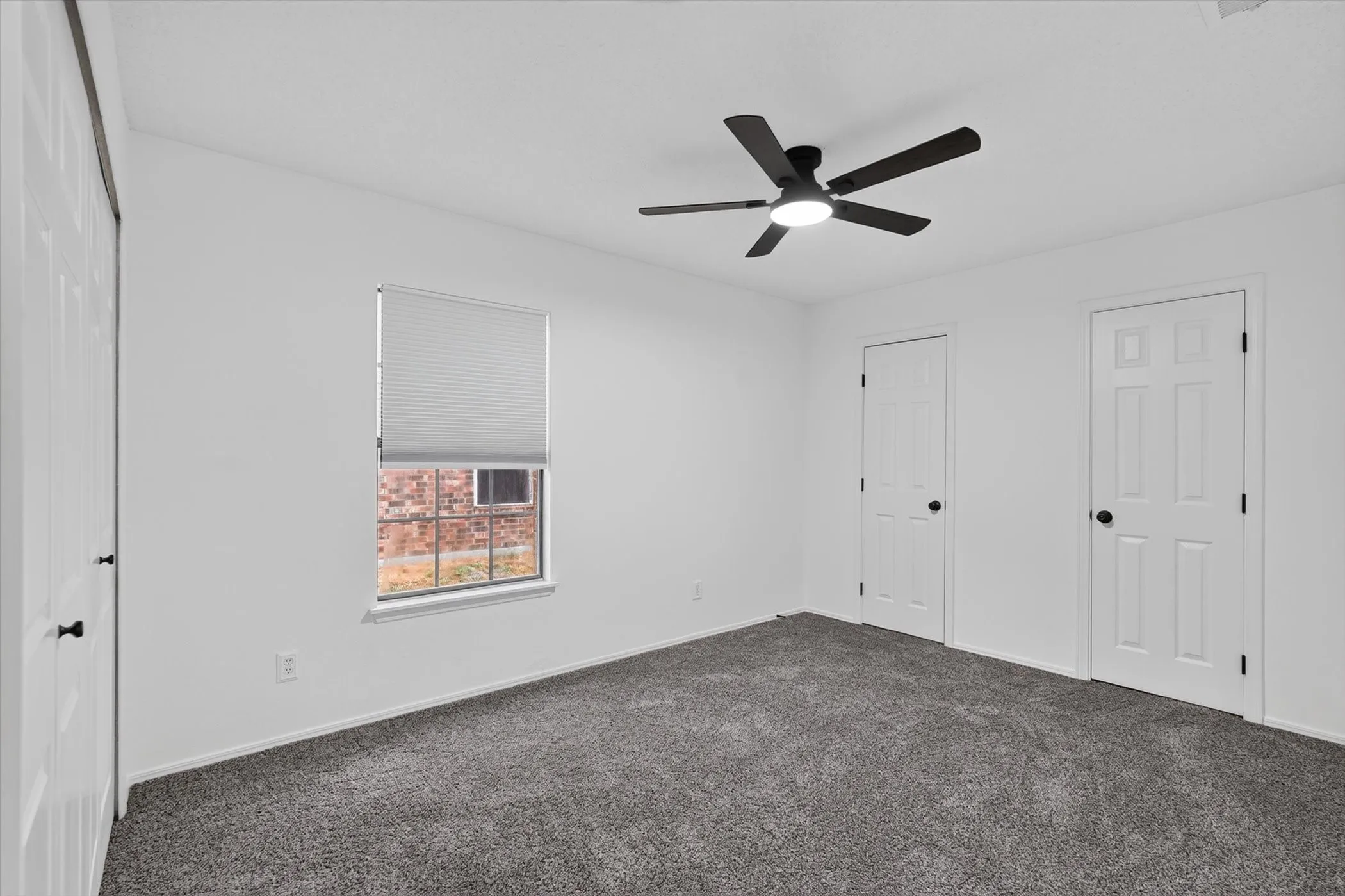 Spacious bedroom with neutral wall finishes, dark gray carpet flooring, ceiling fan with integrated lighting, and multiple white six-panel doors providing access to adjoining spaces