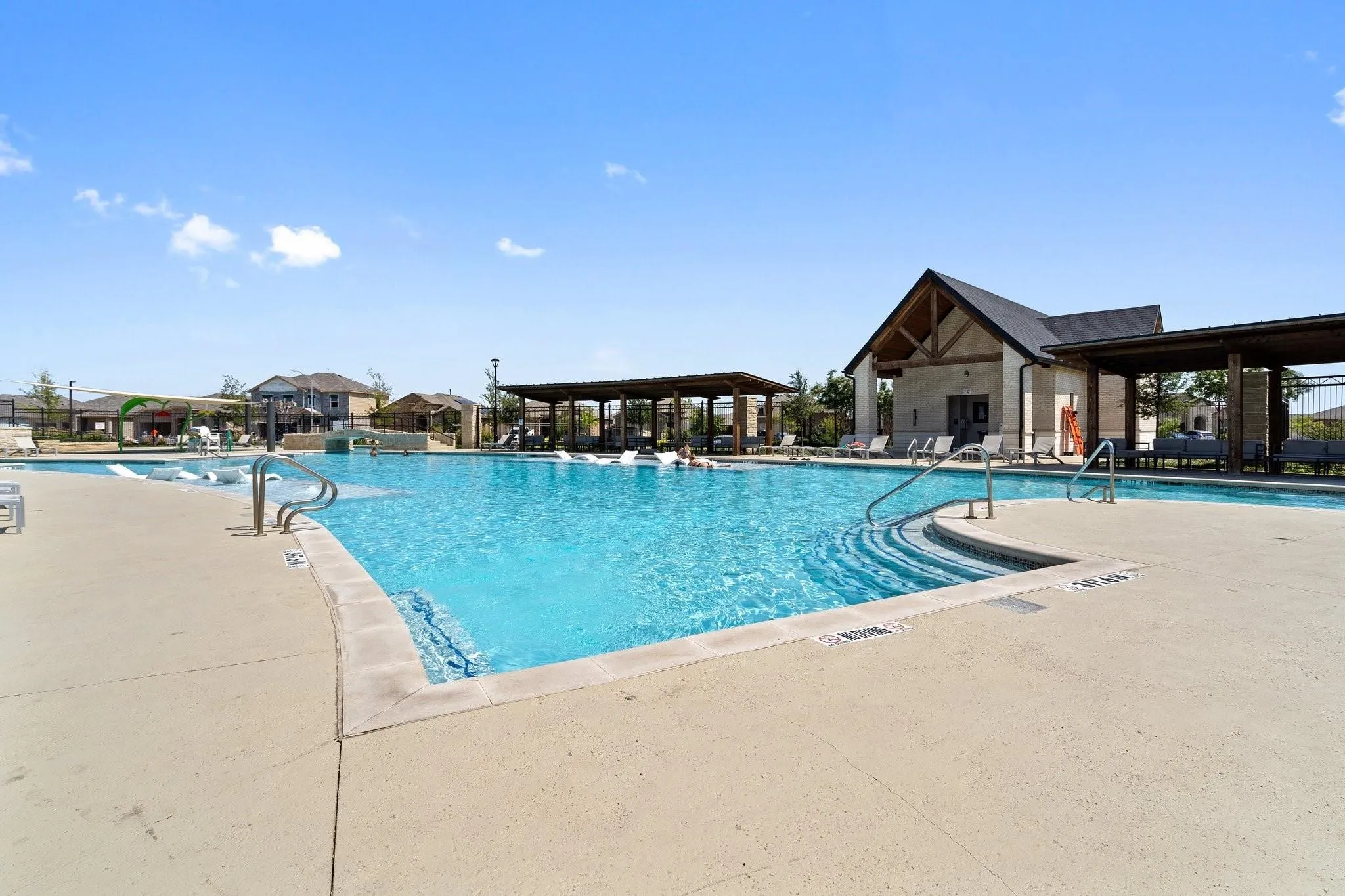 Expansive swimming pool with integrated steps, surrounded by a concrete deck, and shaded pergolas, featuring a central clubhouse structure