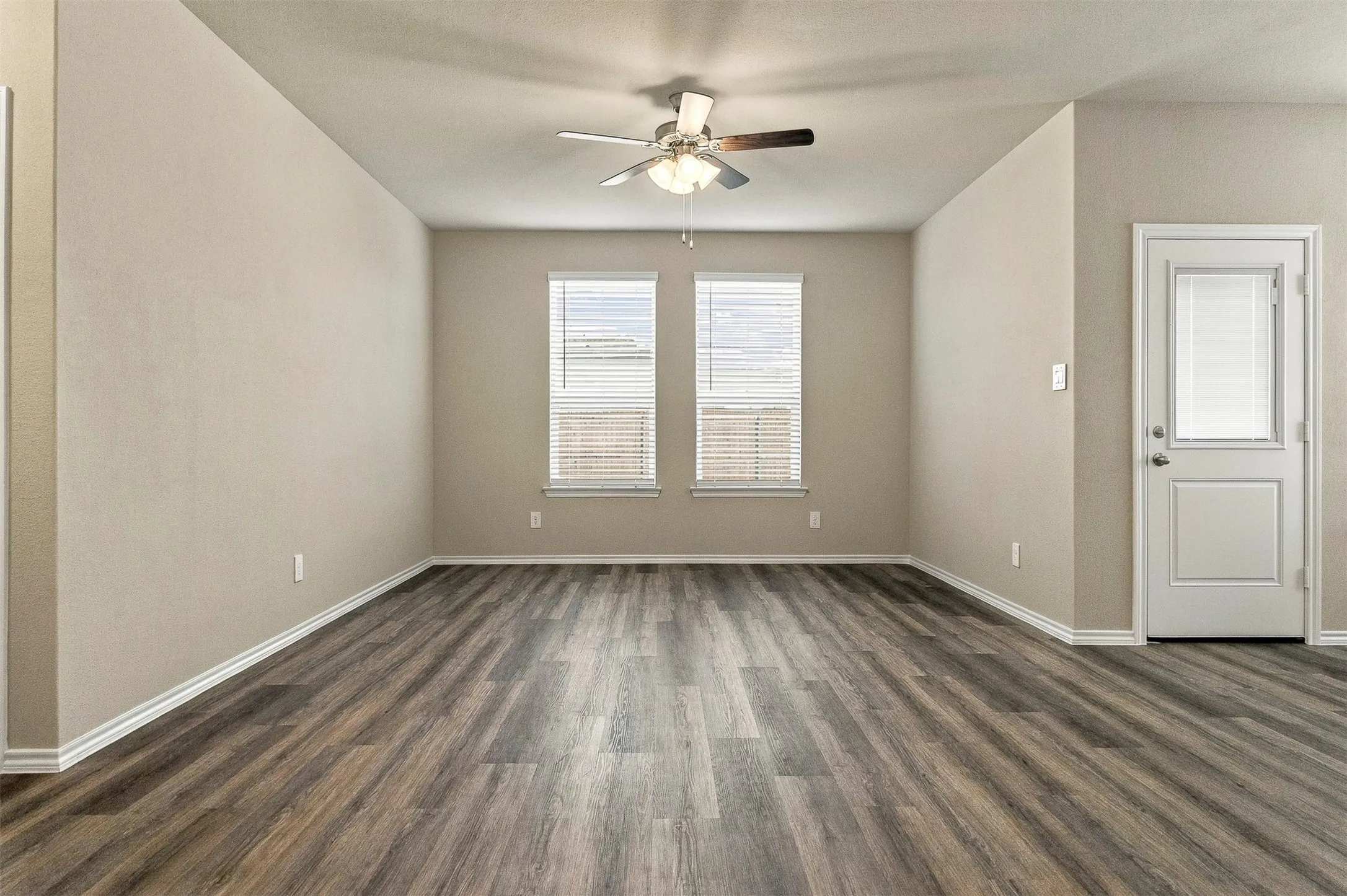 Wood-finish flooring, neutral wall tones, two windows with blinds, a ceiling fan with integrated lighting, and a white paneled door with a glass insert