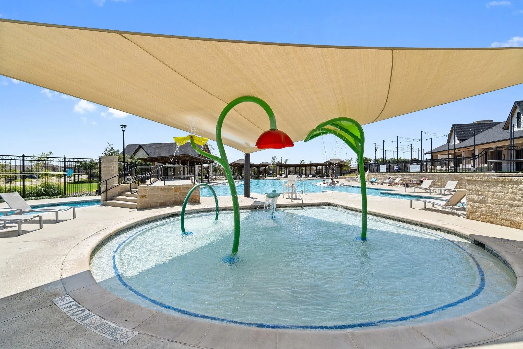 Shaded splash pad with water features, integrated with a large community pool area and adjacent stone-clad structures