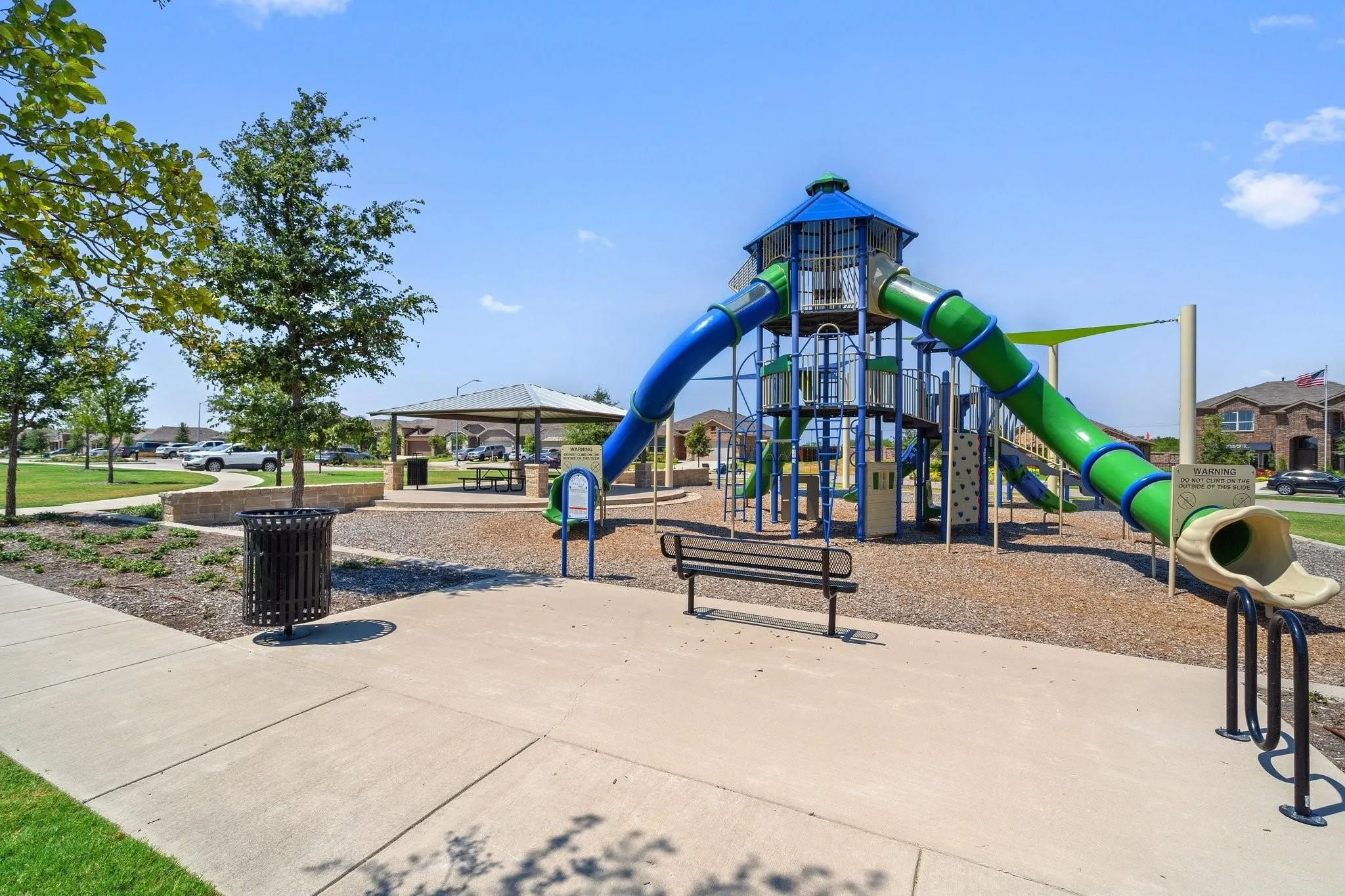Outdoor community recreation area featuring a multi-level playground structure with slides, a covered picnic area, paved pathways, and mature landscaping