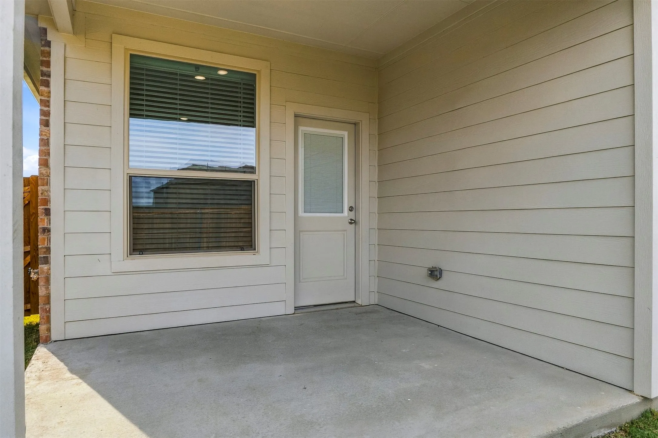 Covered concrete patio with horizontal siding, a single window with blinds, and a white door with a frosted upper pane