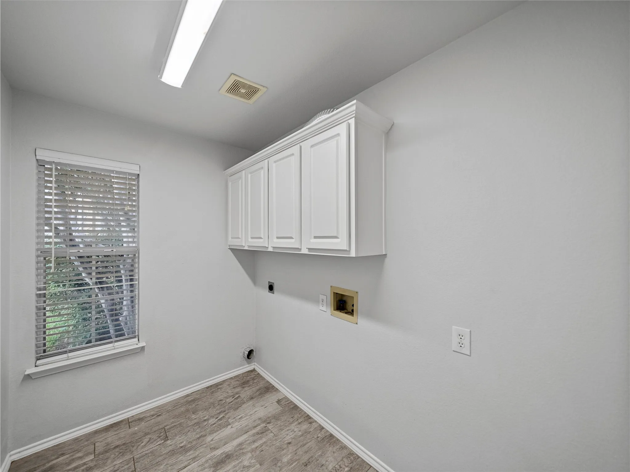Window with blinds, overhead cabinetry, wood-finish flooring, and wall-mounted lighting fixture