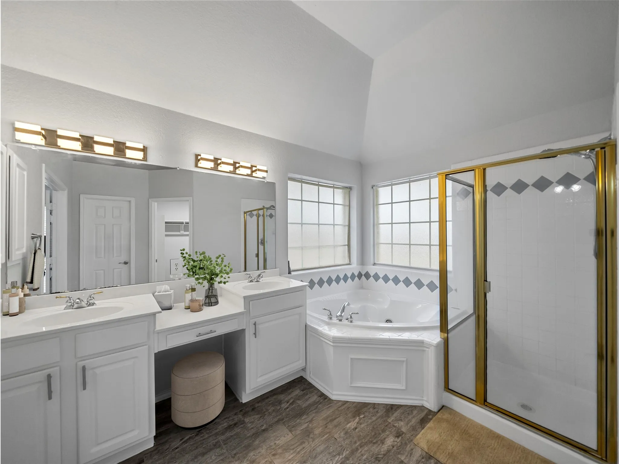 Dual vanity with integrated sinks, built-in vanity area, corner jetted tub, separate shower with brass-finish framing, and wood-finish flooring