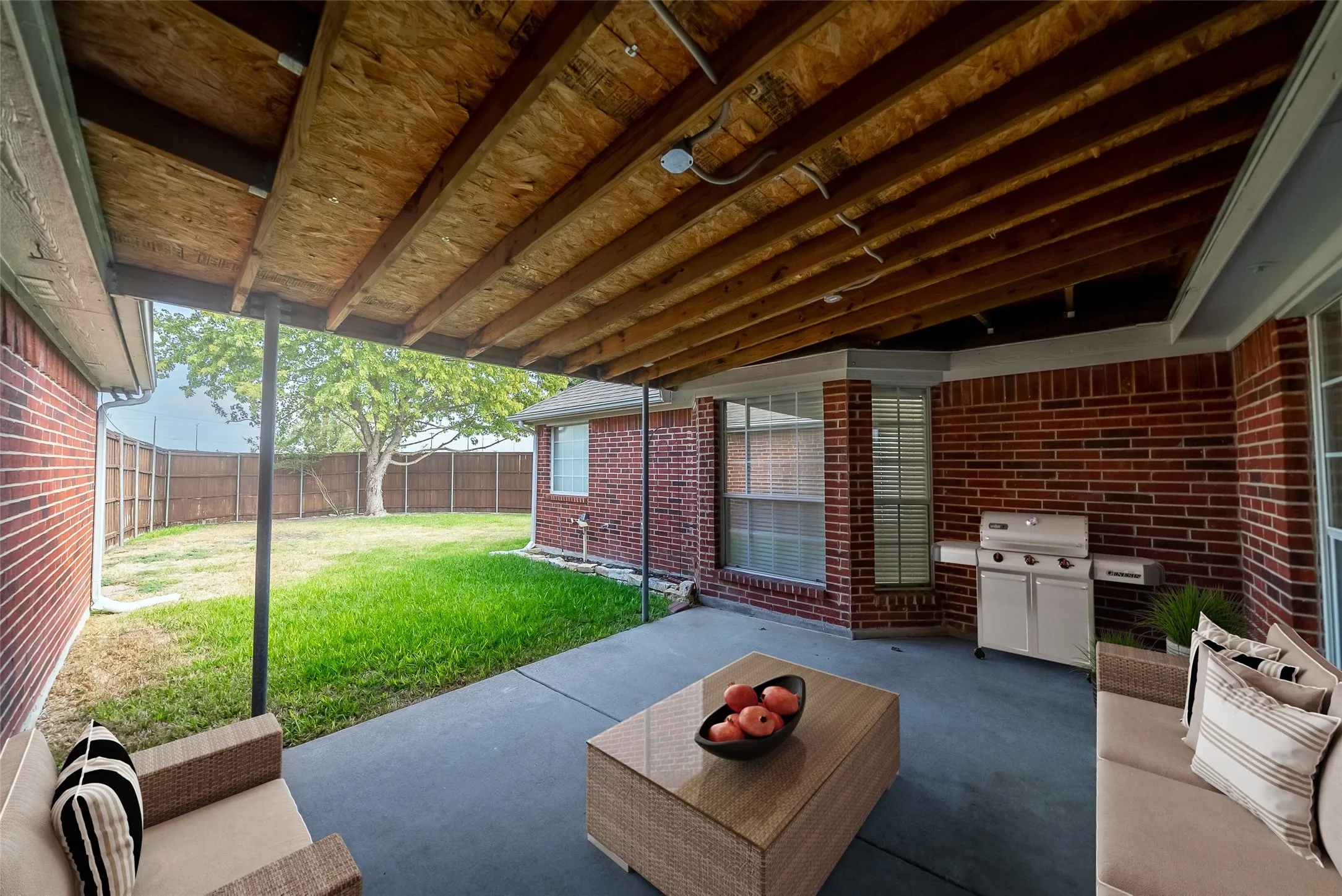 Covered patio with a concrete surface and exposed wood rafters, featuring a brick exterior wall with a bay window, and a fenced grassy yard