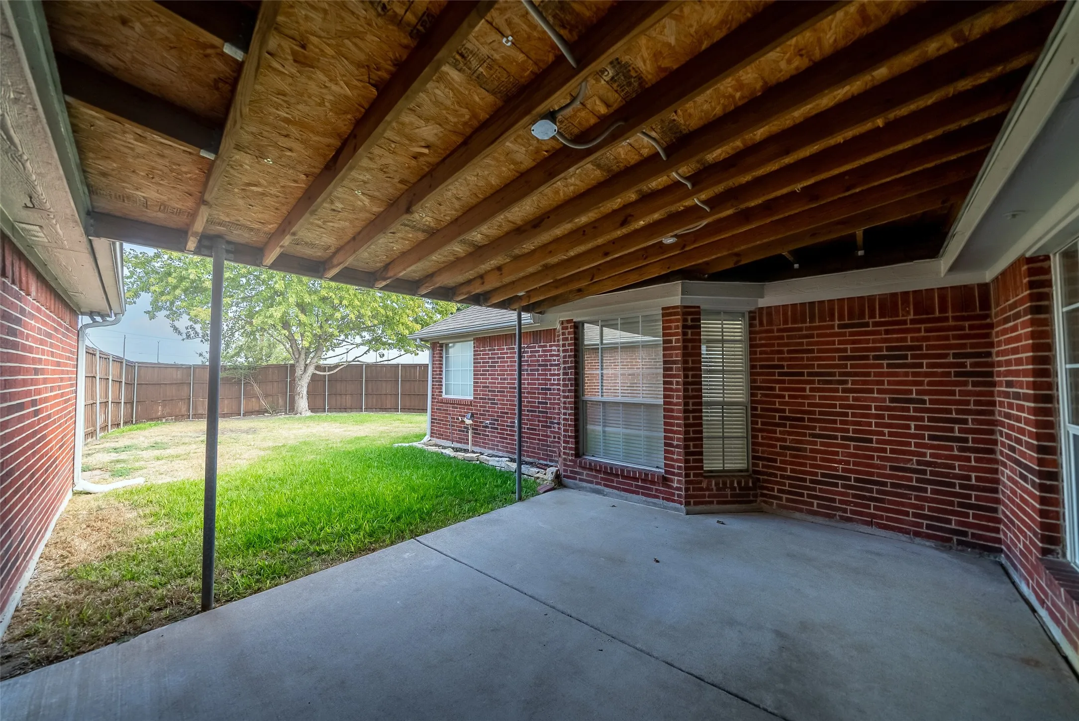 Covered patio with concrete flooring, brick exterior, and a fenced yard with mature trees