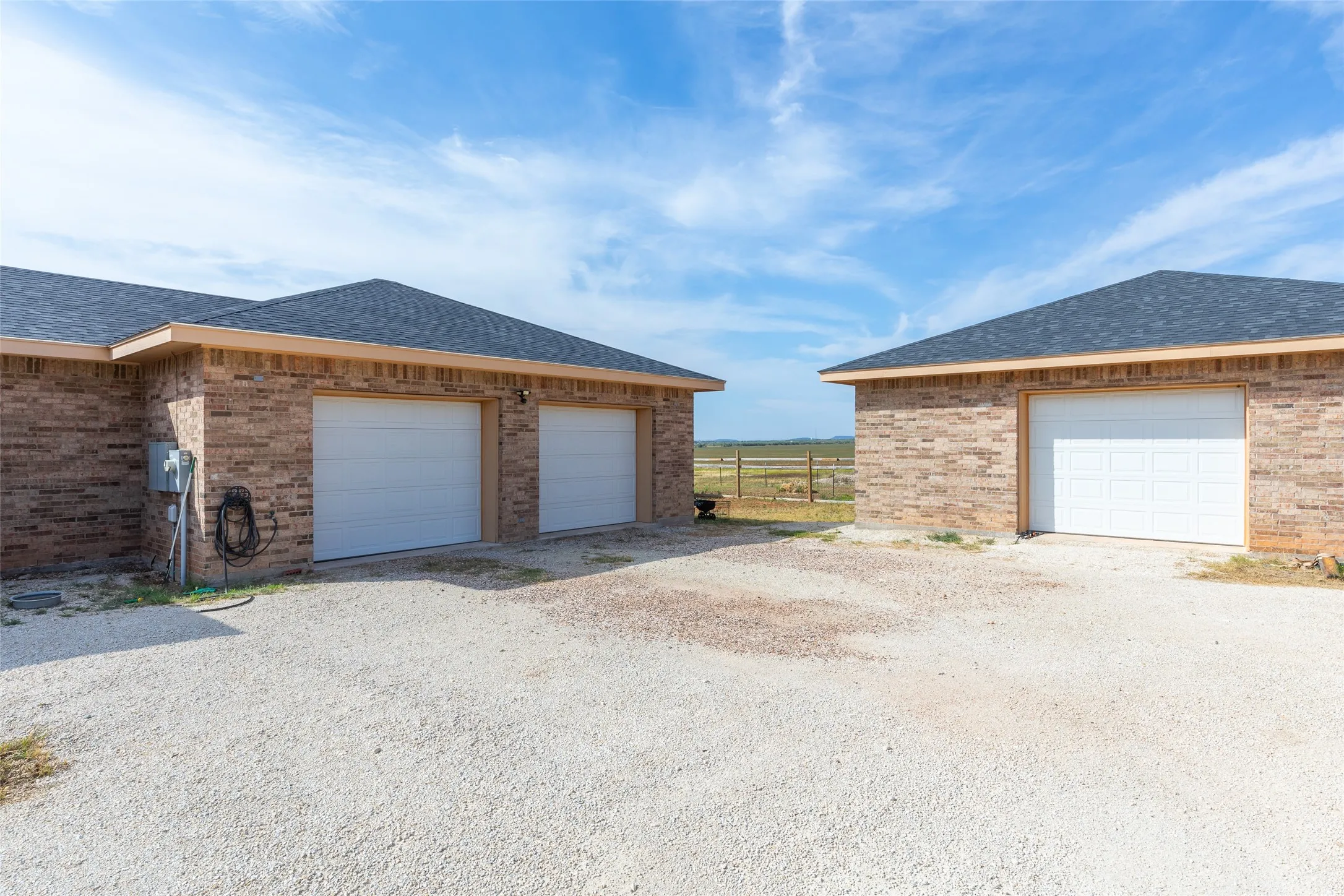 Two brick structures with dark shingle roofs, featuring multiple white garage doors and a gravel driveway