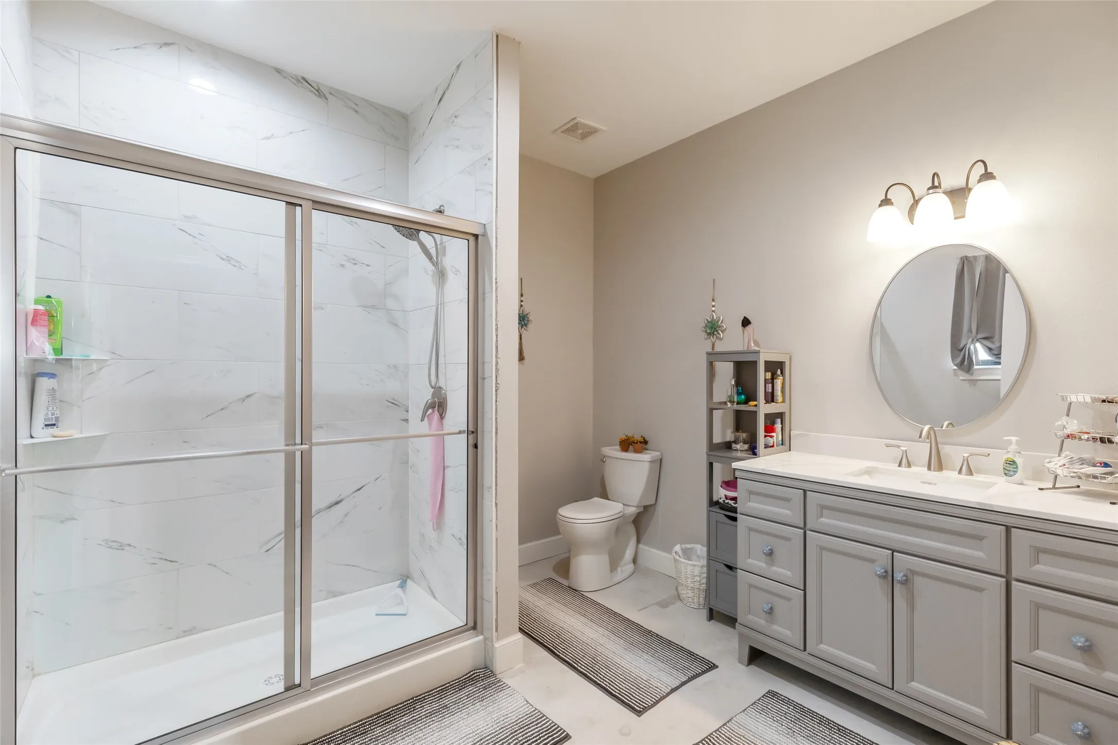 Spacious bathroom featuring a double vanity with light gray cabinetry and white countertops, a round mirror, and a glass-enclosed shower with white tiled walls